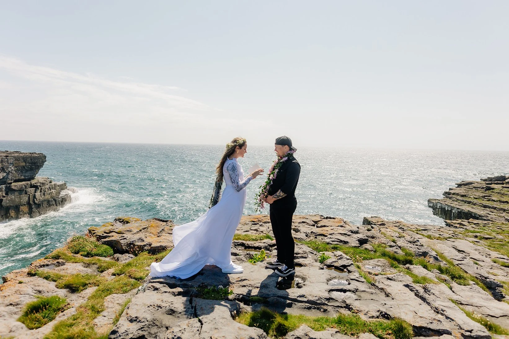 A bride and groom exchanging wedding rings on a rocky cliff by the ocean. The bride is in a white wedding dress with lace sleeves, and the groom is in a black suit with a floral lei and a backward cap. The background features the sea, sky, and rugged coastline.