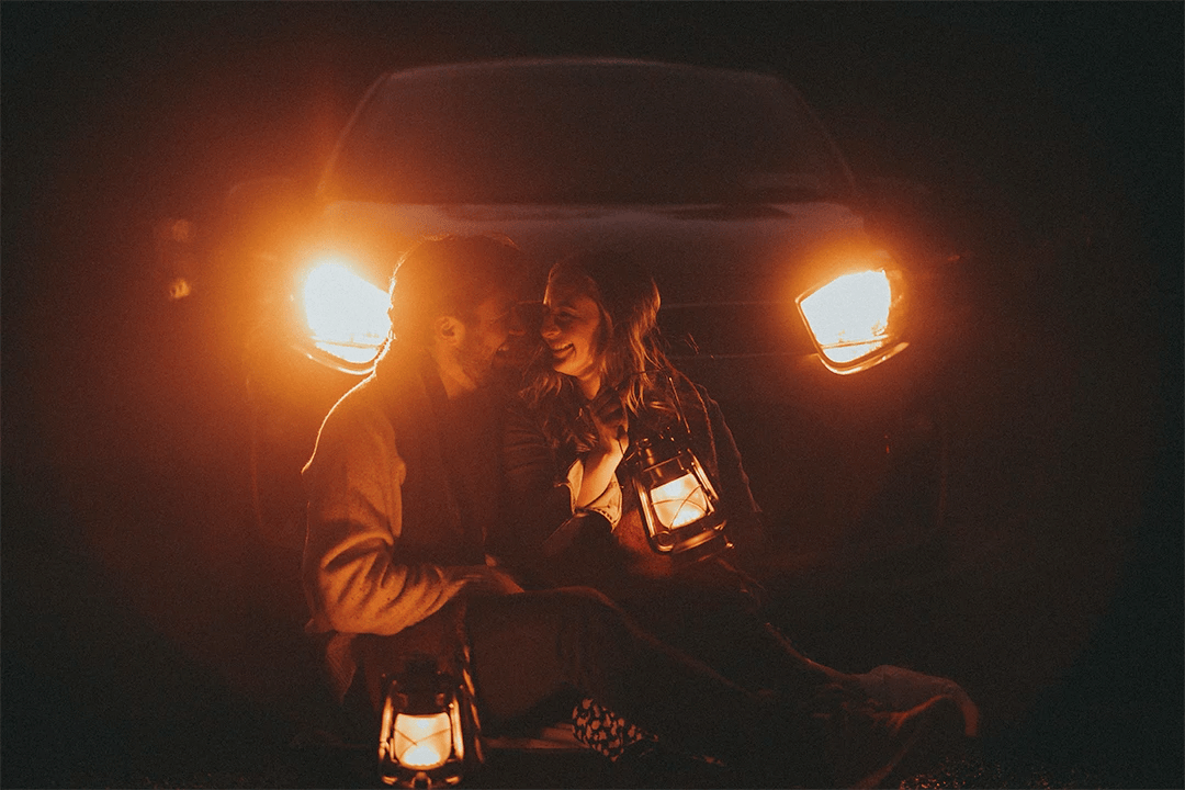 A couple sitting closely in front of a car with headlights on at night, holding lanterns and smiling while enjoying each other’s company during their adventure couple shoot.