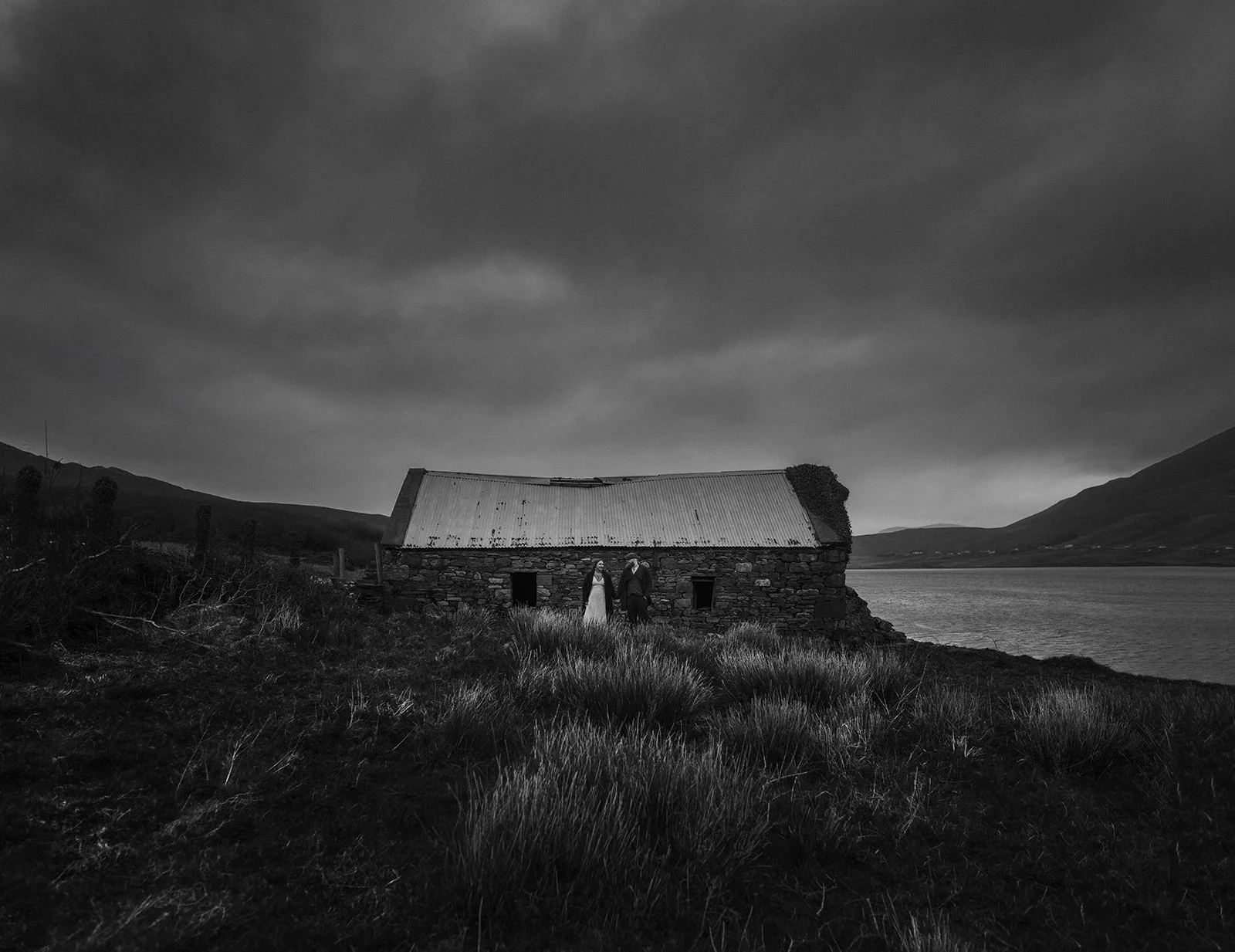 Black and white photo of a couple dressed formally standing outside an old stone house with a metal roof, near a lake with mountains in the background under a cloudy sky.