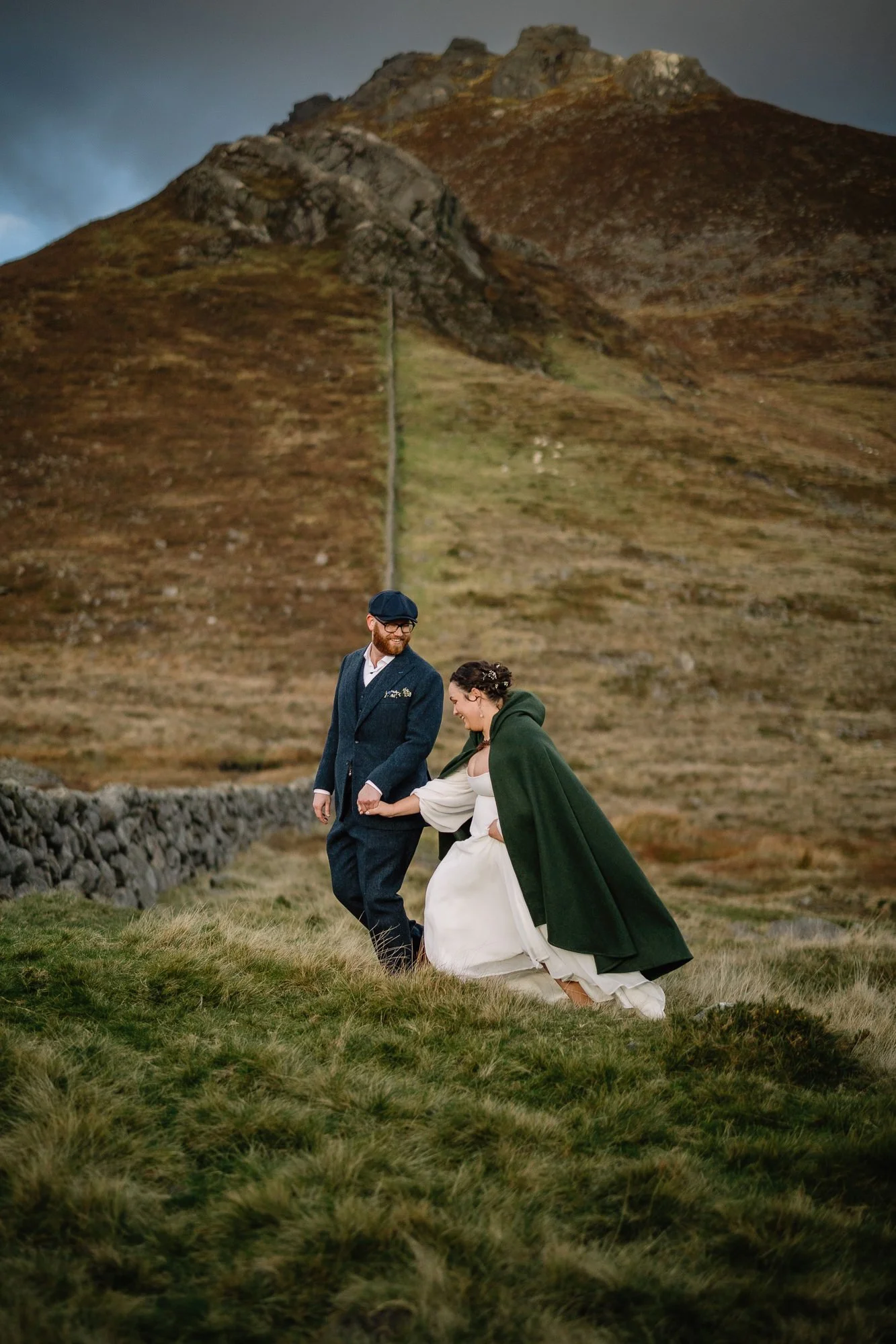 A couple dressed in wedding attire walking outdoors in a grassy area with a stone wall and a large brown hill in the background.