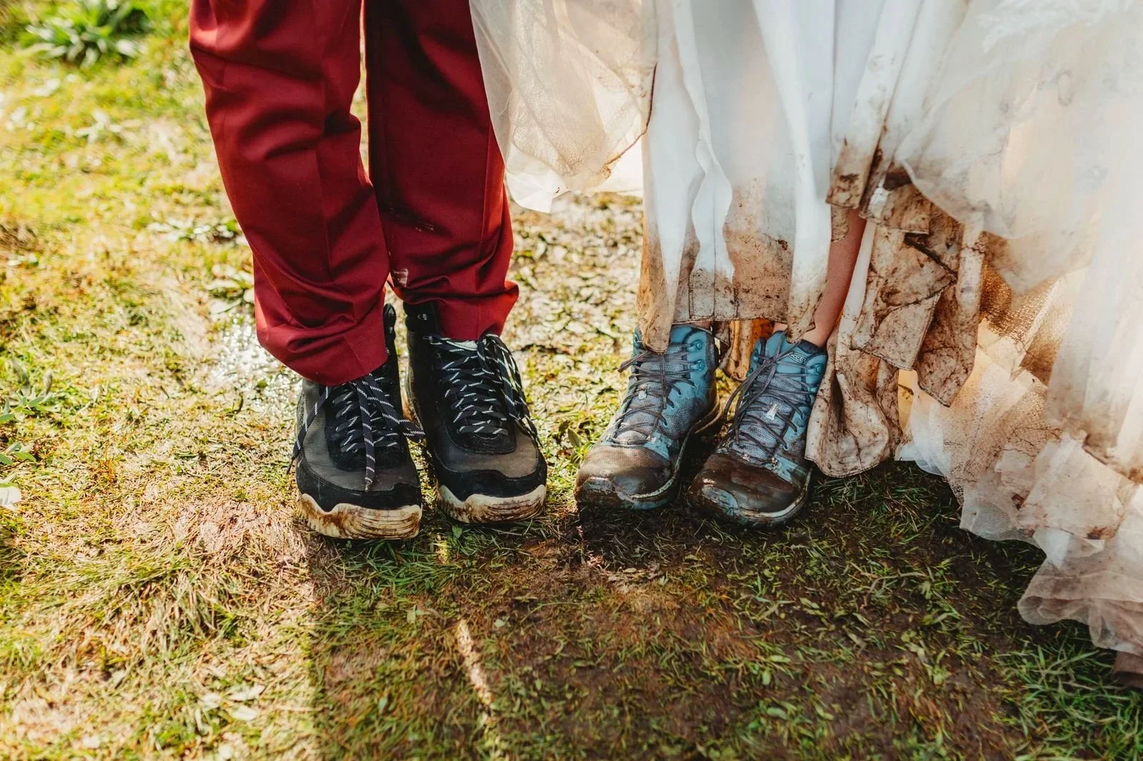 Close-up of muddy hiking shoes on muddy and grassy outdoor terrain, with parts of two people visible wearing hiking gear.