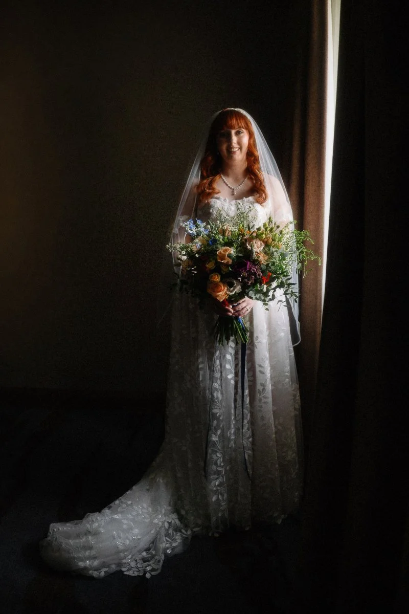 A bride with red hair smiling, wearing a white lace wedding dress and veil, holding a colorful bouquet of flowers, standing near a window with dark curtains.