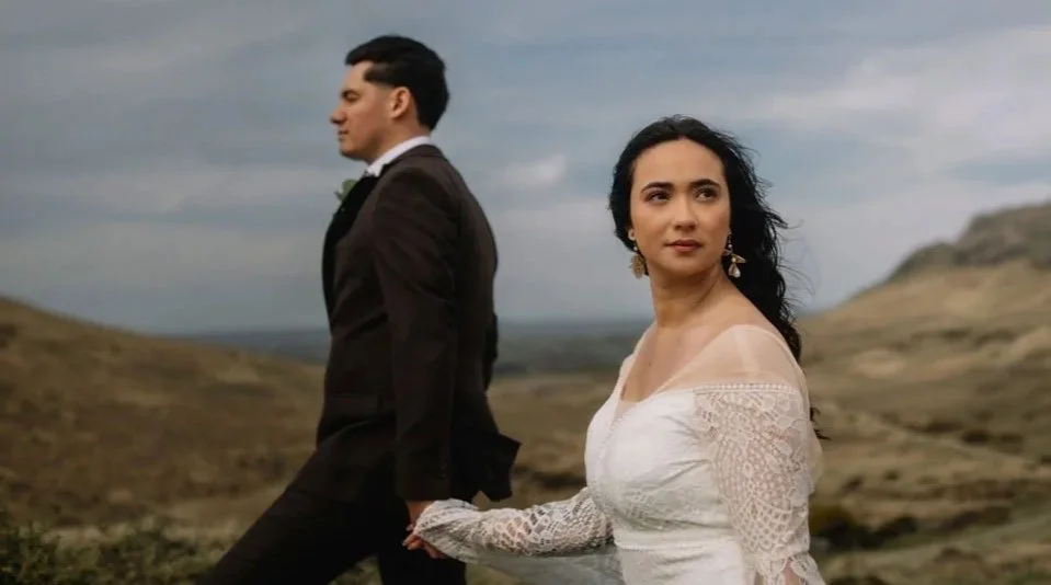 A couple holding hands outdoors in a rocky landscape, the woman looking into the distance and the man facing away.