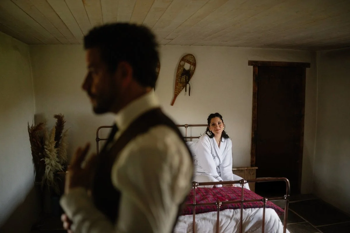 A woman sitting on a bed in a room, looking at a man who is out of focus in the foreground. The woman is wearing a white robe and smiling. The room has a wooden ceiling, a rustic door, a vintage sled hanging on the wall, and dried plants in a vase.