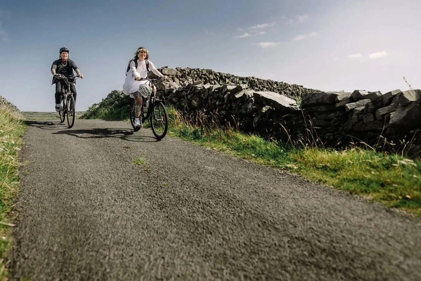 A Couple riding bicycles on a gravel road beside a low stone wall, with a clear sky overhead.