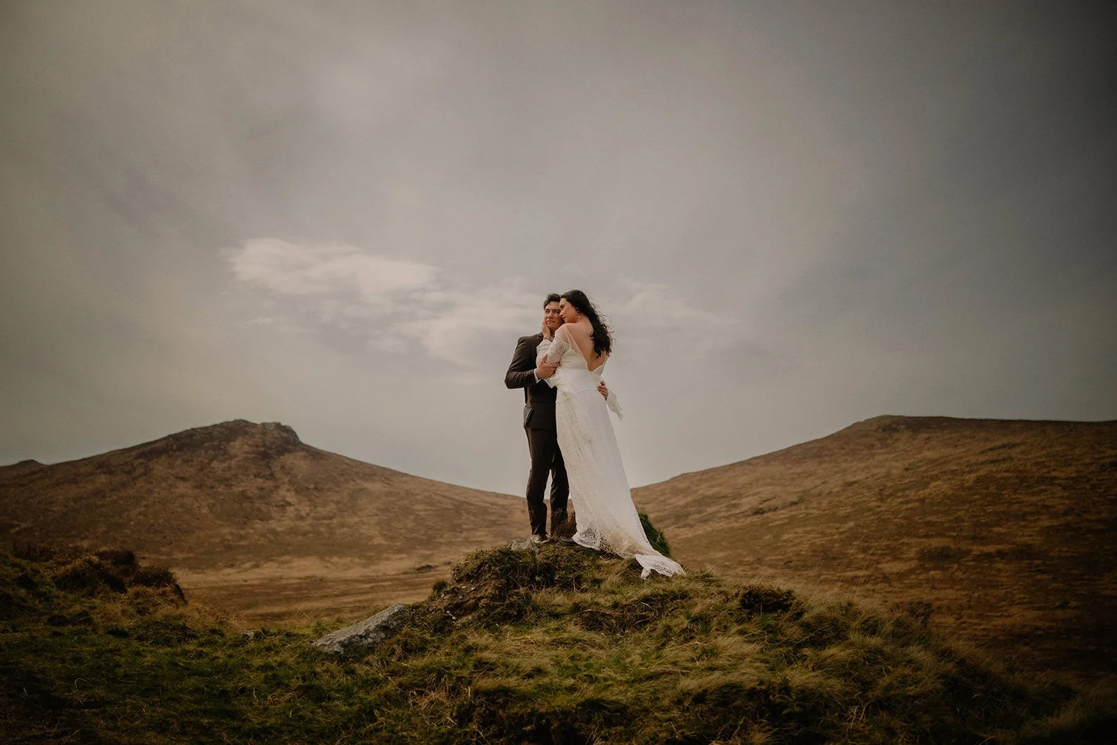 A bride and groom stand embracing on a small hilltop in an open landscape with rolling hills and cloudy sky in the background.