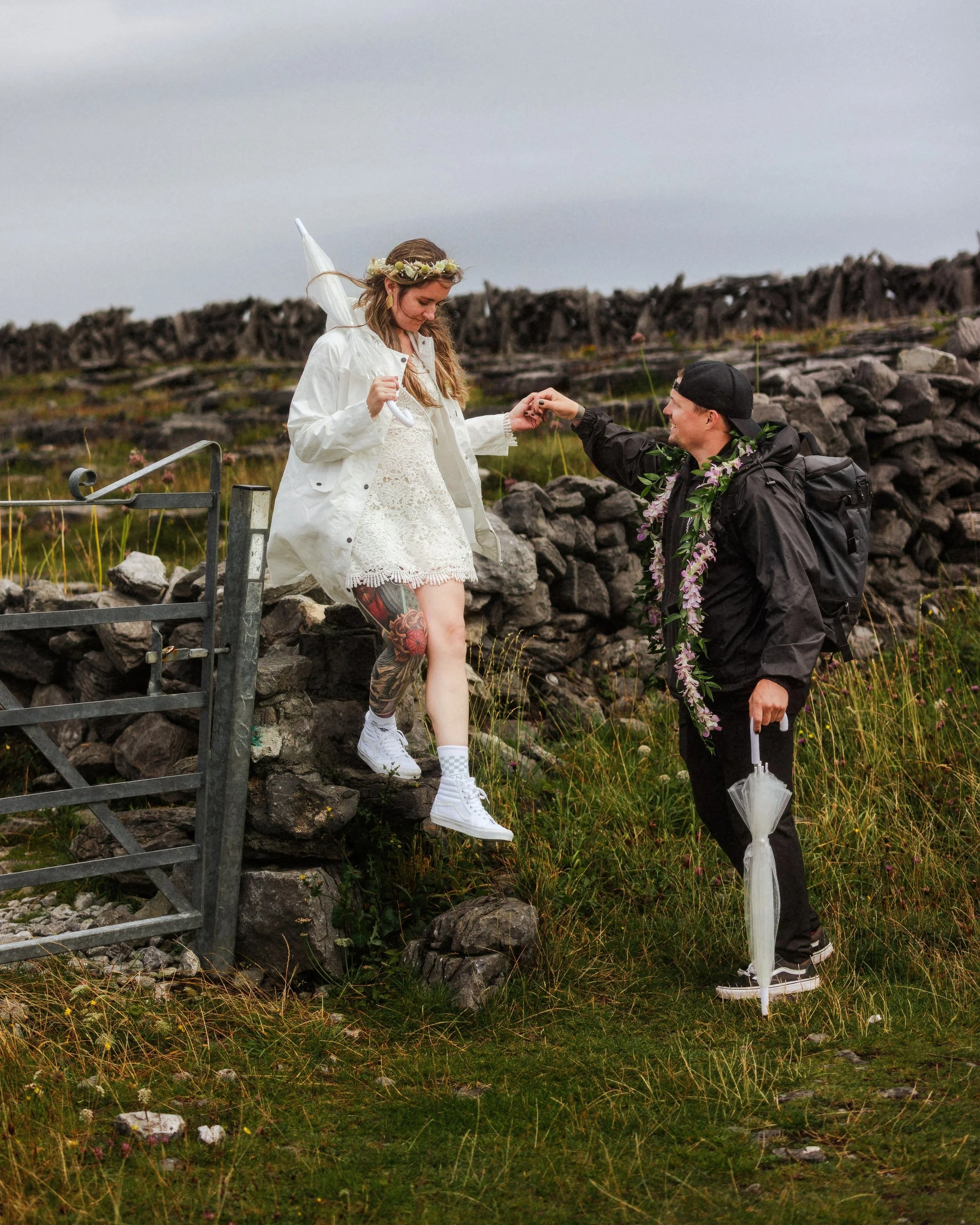 A couple is outdoors, with the woman wearing a white lace dress, white sneakers, and a flower crown, stepping down a rocky stone wall, holding an umbrella, while the man, dressed in black with a black cap and backpack, offers her a small item, and is