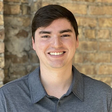 Smiling person in front of brick wall, wearing a gray shirt.
