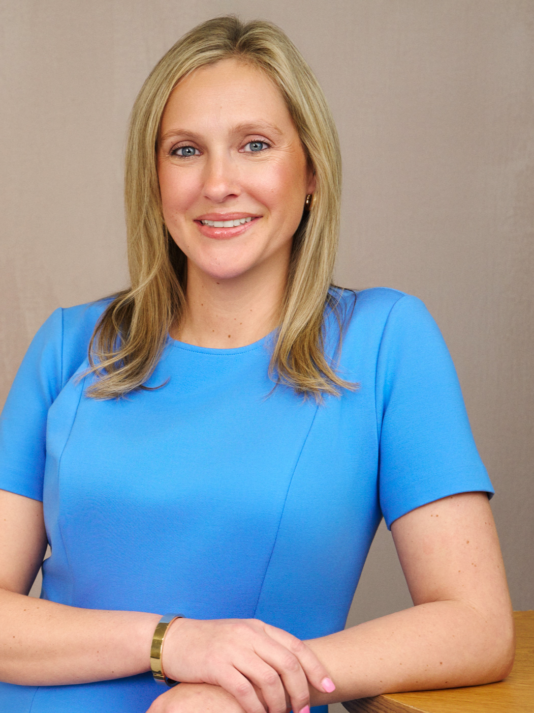 A woman with blonde hair and blue eyes smiling, sitting at a table, wearing a bright blue dress and a gold bracelet, in front of a neutral background.