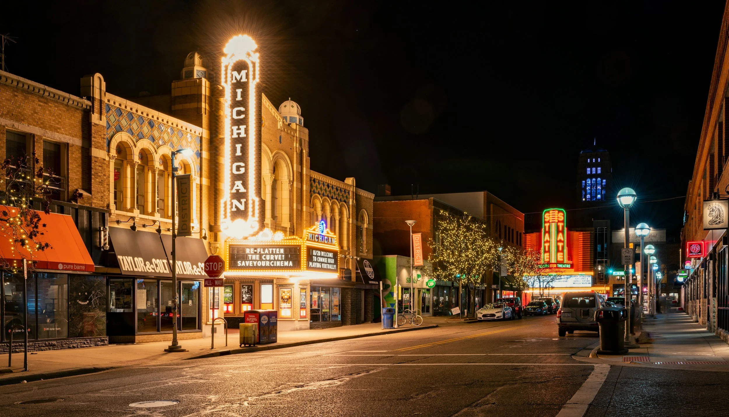 Night view of a downtown street with buildings decorated with colorful lights, illuminated signs, and Christmas lighting, including a marquee for the Michigan Theatre and a large neon 'Stature Theatre' sign.