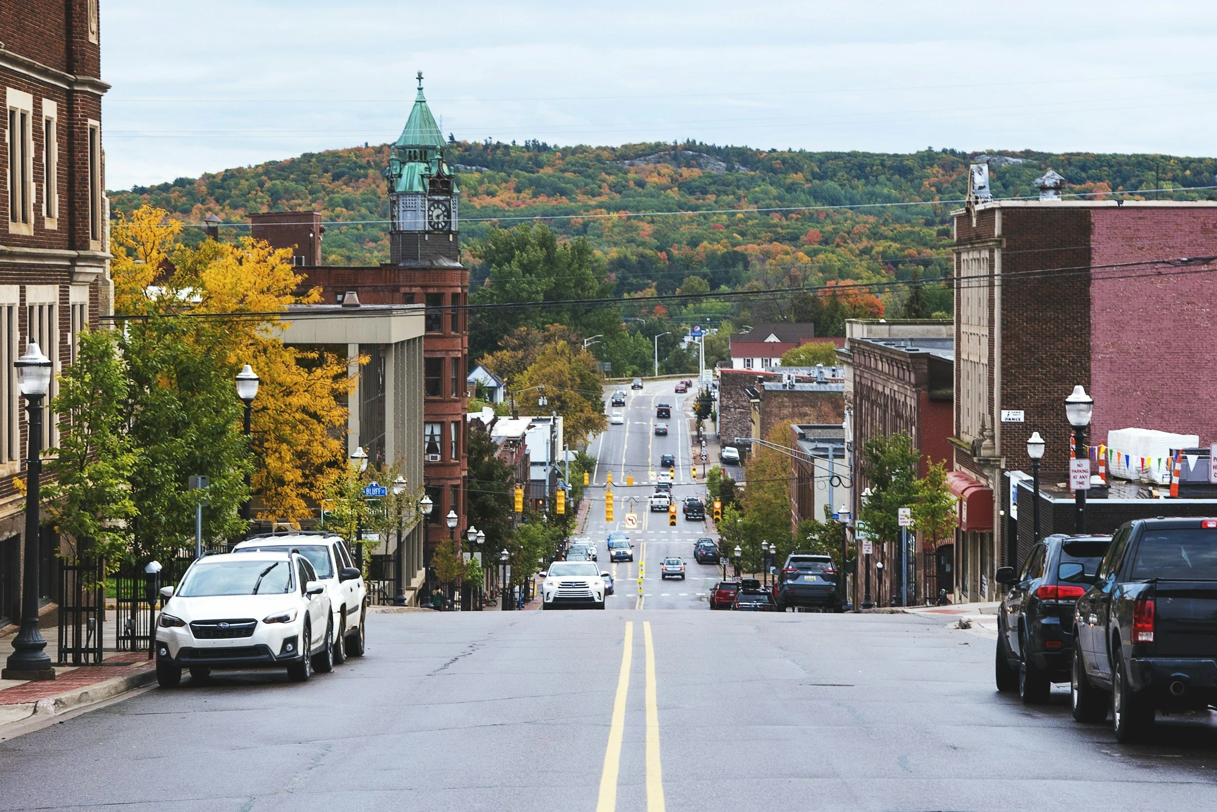 Downtown Michigan street view with cars parked along the sides, buildings with brick facades, streetlamps, and a church with a green steeple in the background, overlooking a hill covered in trees with fall foliage.
