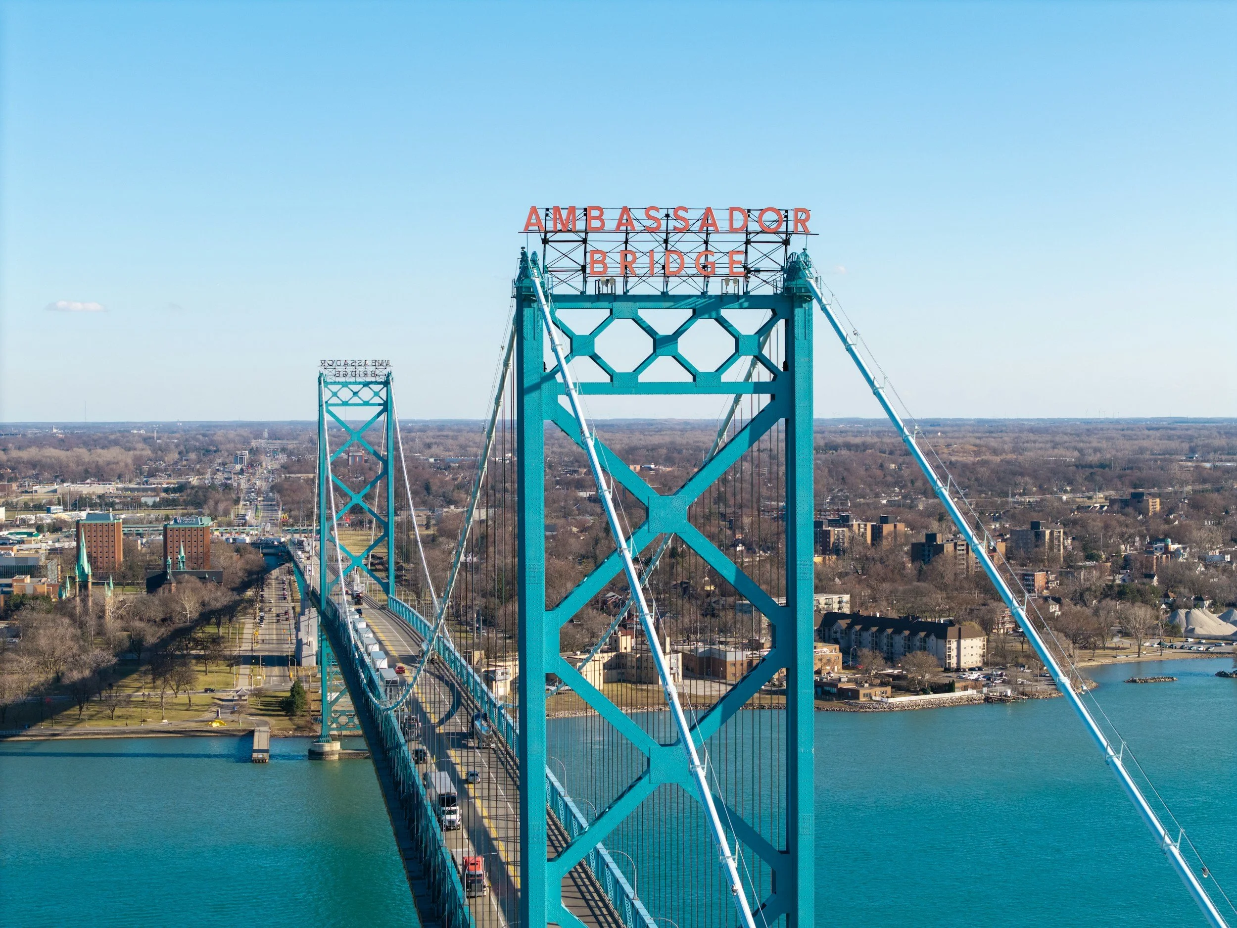 A suspension bridge painted blue with a sign spelling 'AMBASSADOR BRIDGE' in red letters at the top, spanning over a body of water with a cityscape and roads in the background under a clear blue sky.