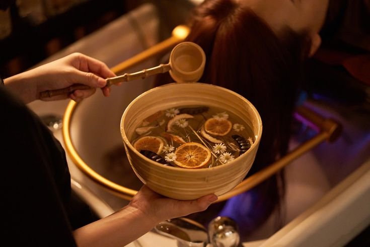 Person holding a bowl of herbal tea with slices of dried citrus and flowers, with another person in the background.