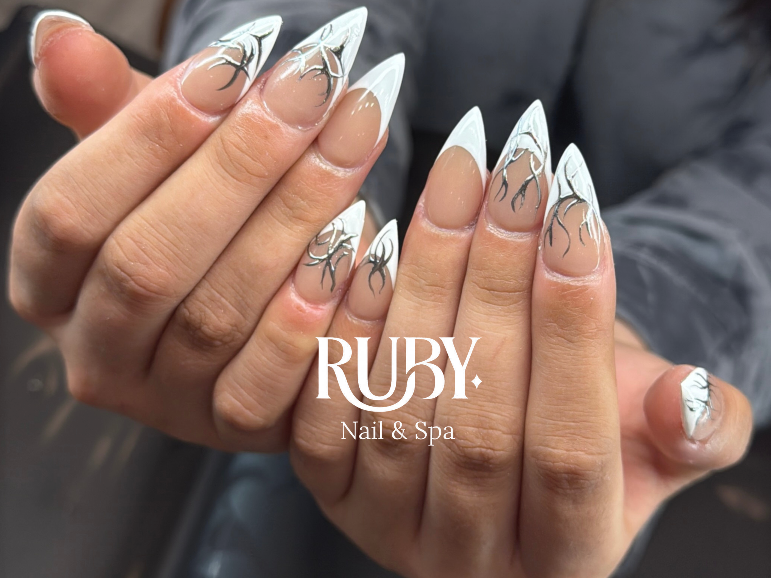 Close-up of a person’s hand with long, almond-shaped nails featuring white and black abstract designs, held against a dark background. The image includes a logo for 'Ruby Nail & Spa'.