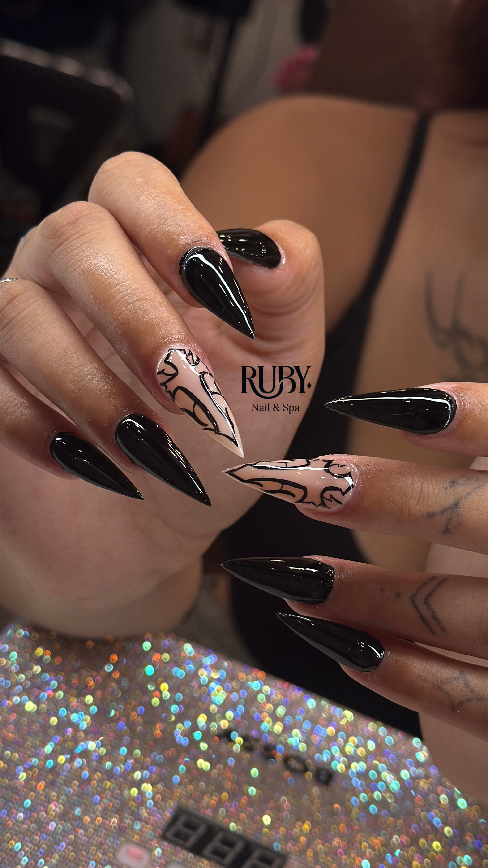 Close-up of a hand with long, stiletto-shaped nails painted black and decorated with nude color and black abstract line art. The background shows a woman with a tattoo on her arm, wearing a black strap top, and a sparkly, multicolored surface.