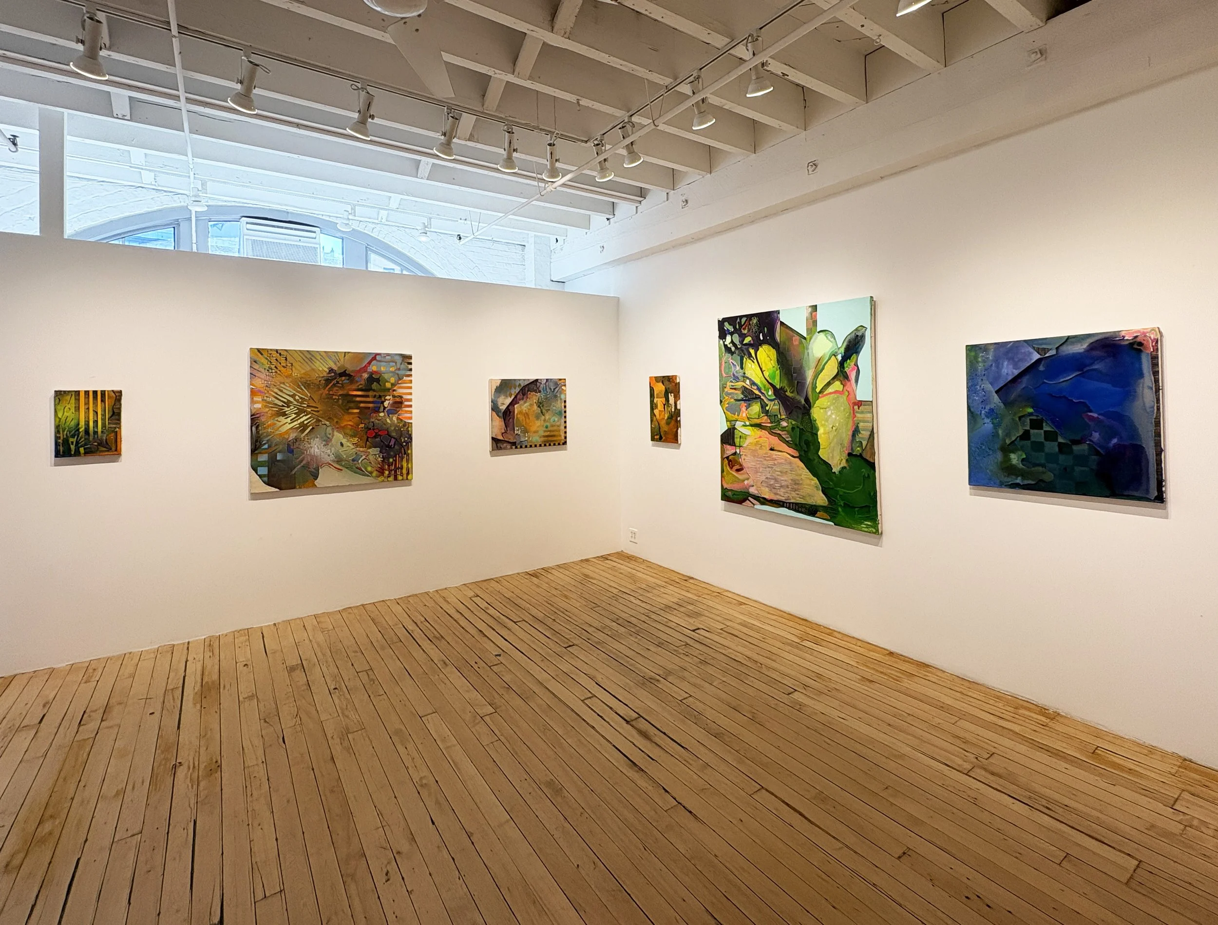 Art gallery with colorful abstract paintings hanging on white walls, wooden floor, and a high ceiling with track lighting and a large window.