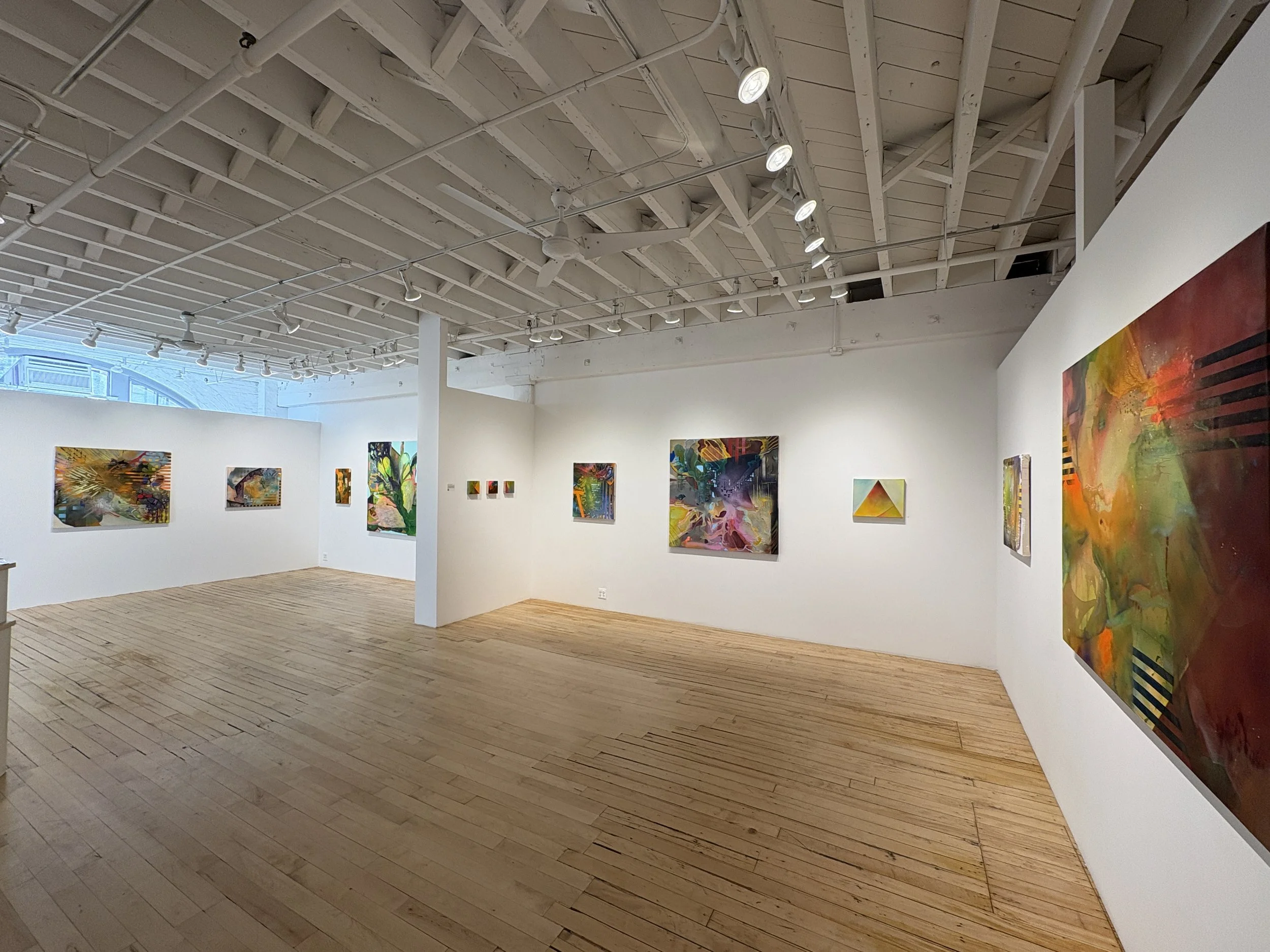 Interior of an art gallery with white walls and wooden flooring, displaying various colorful abstract paintings hanging on the walls.