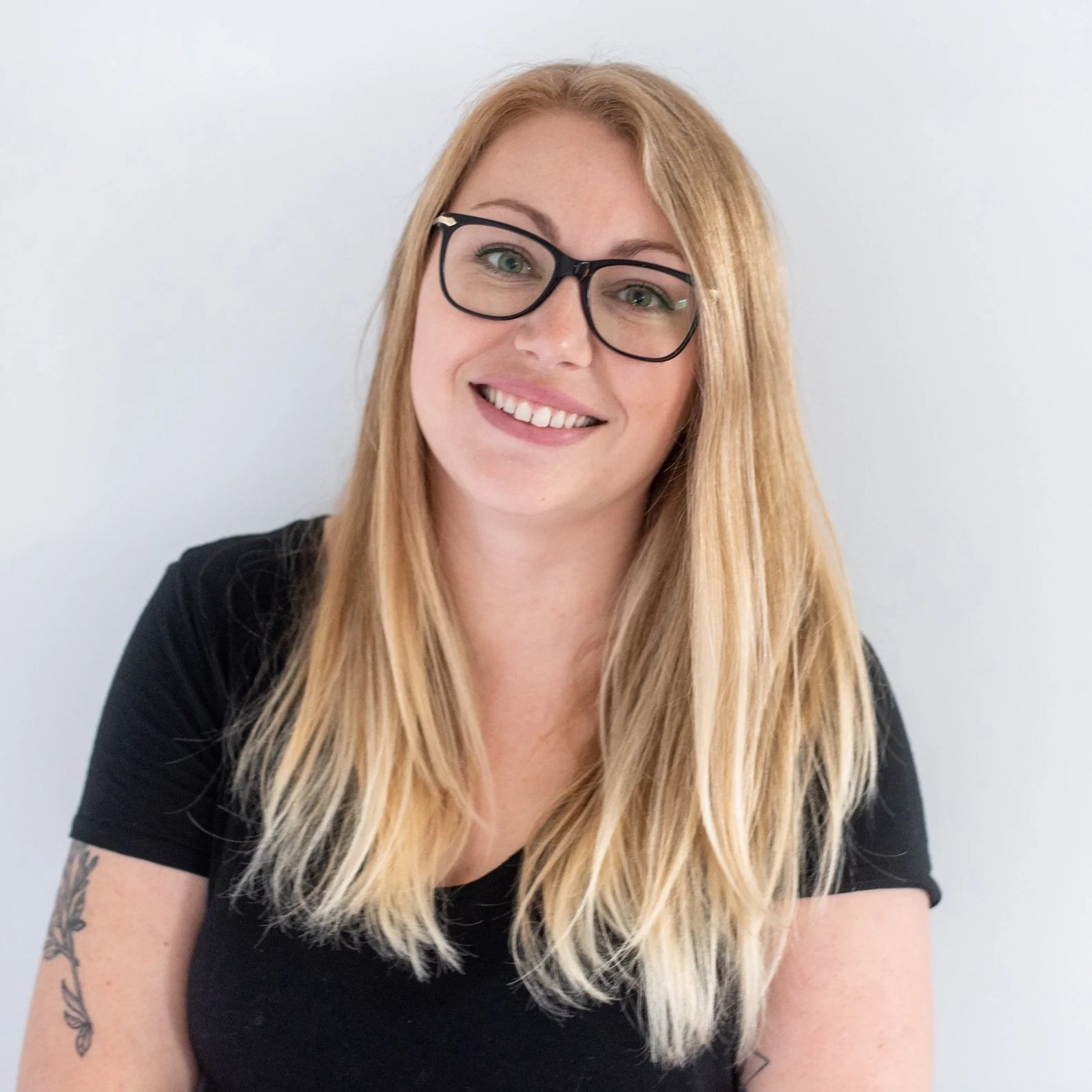 Woman with glasses and long blonde hair smiling, wearing a black shirt against a plain background.