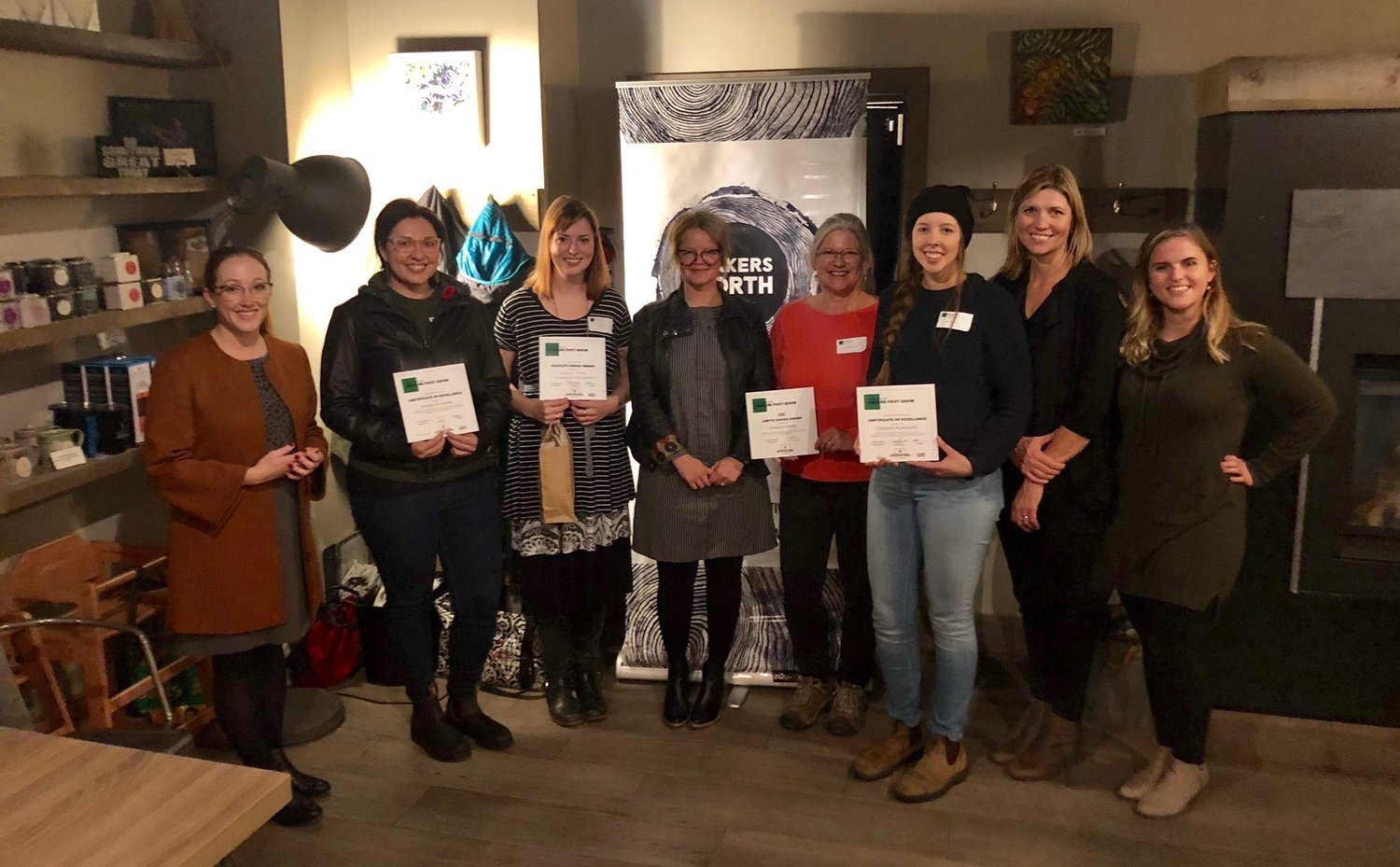 Group photo of eight women standing indoors holding certificates, smiling; background includes wall art and shelves with items.