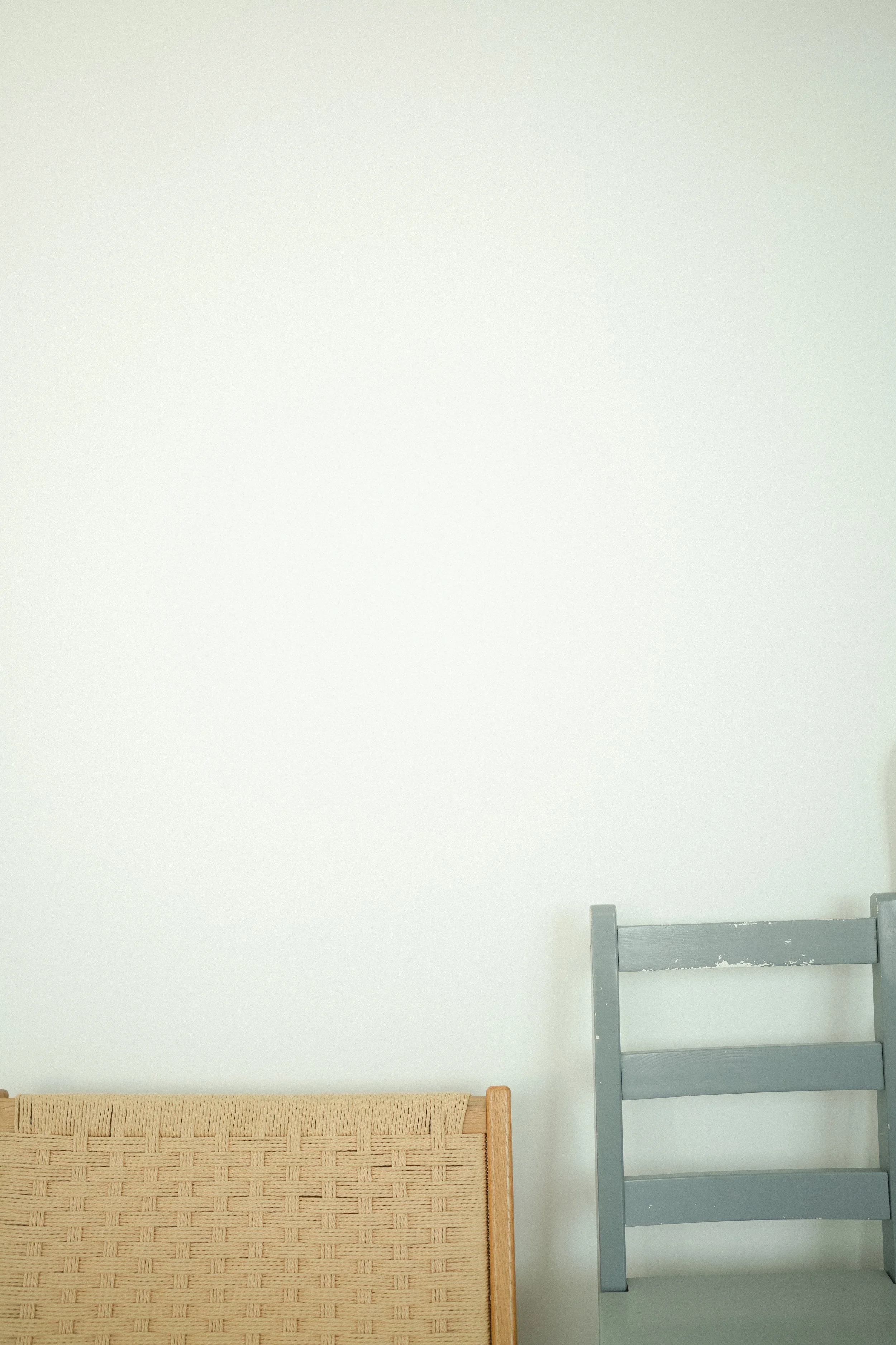 Two chairs, one with a woven wooden backrest and the other painted gray, are against a white wall.