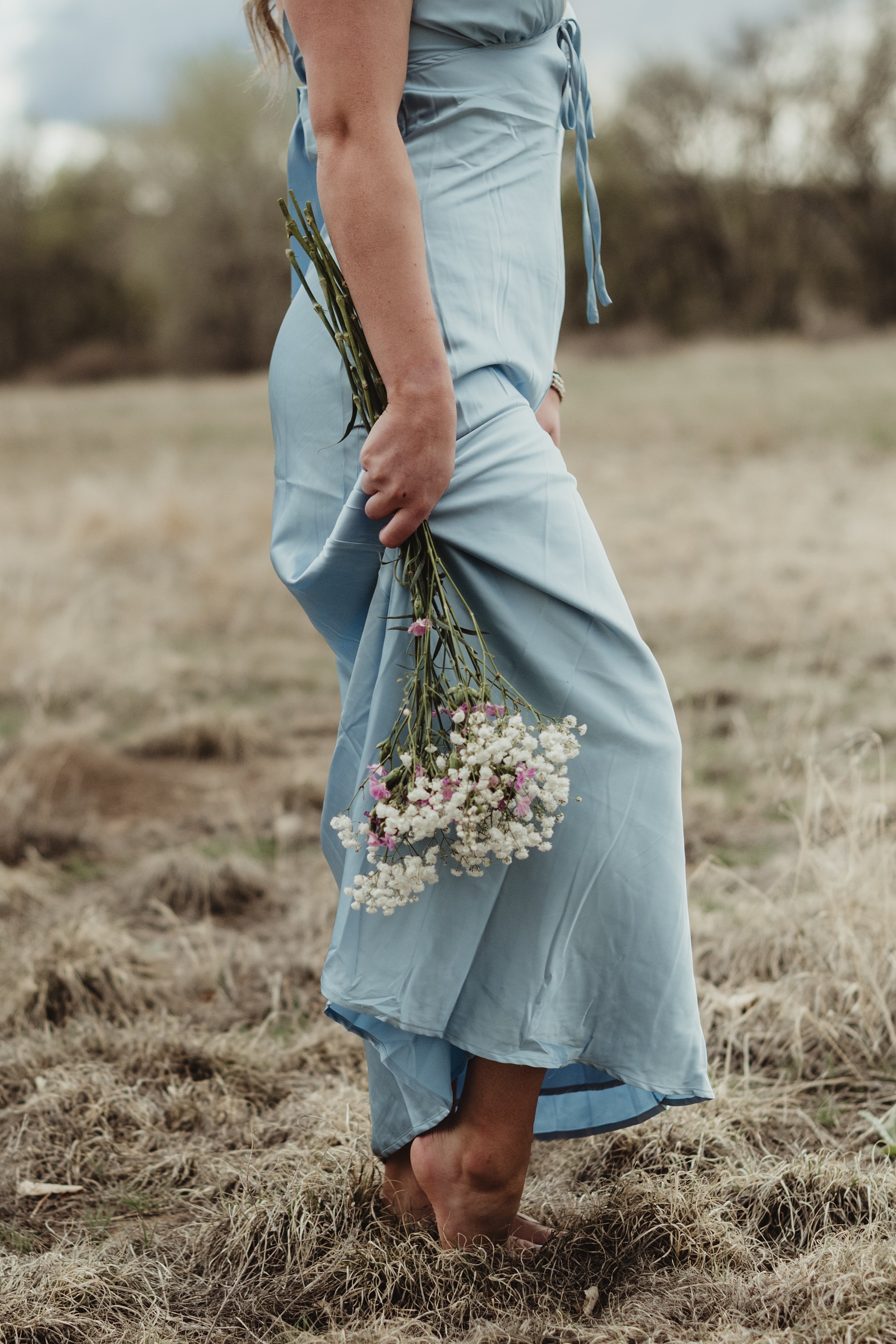 A woman standing outdoors in a field of dry grass, holding a bouquet of white and pink flowers, wearing a light blue dress.