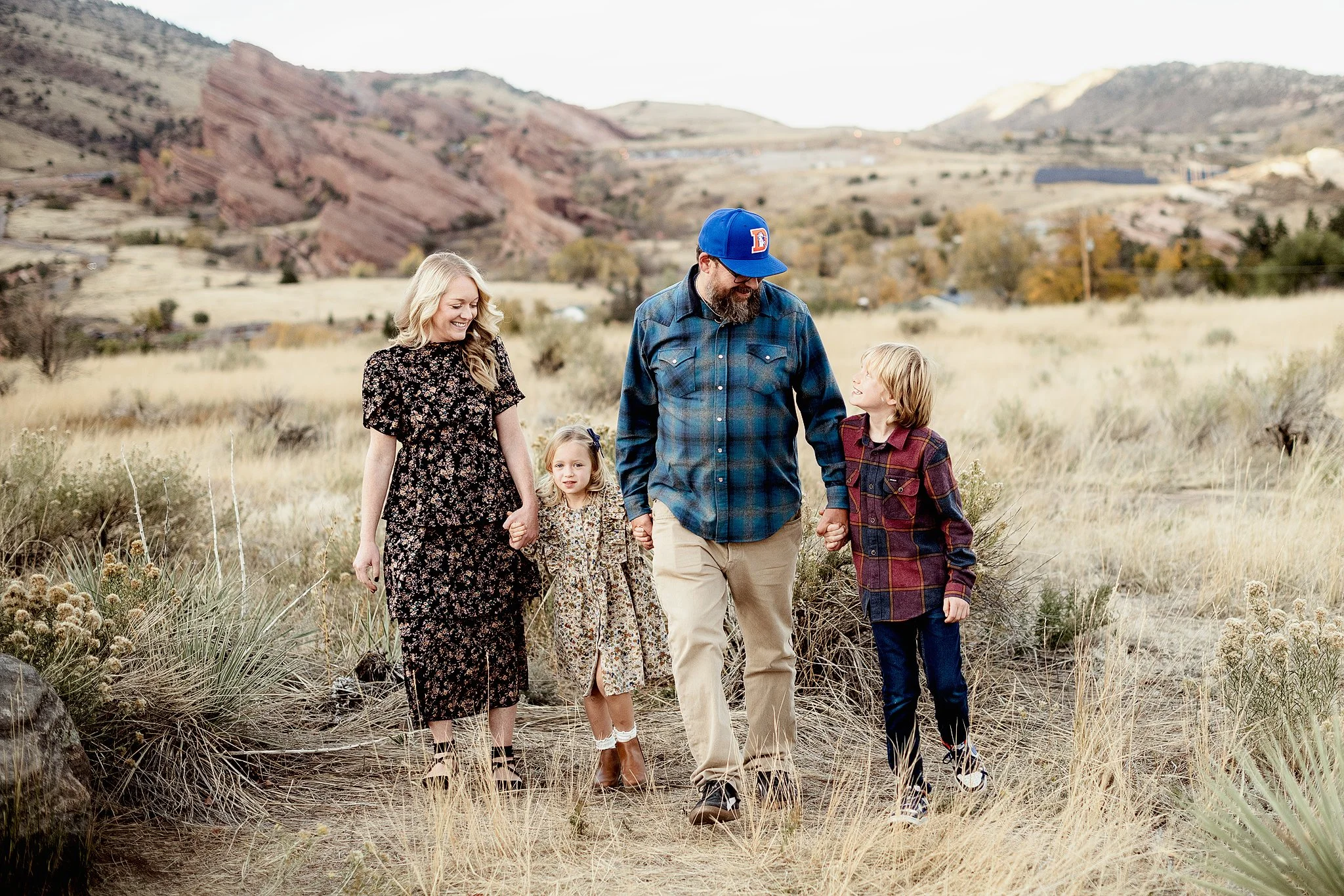 A family of five walking outdoors in a grassy field with mountains in the background. The family includes a woman, a man, two young girls, and a teenage boy, all smiling and holding hands.