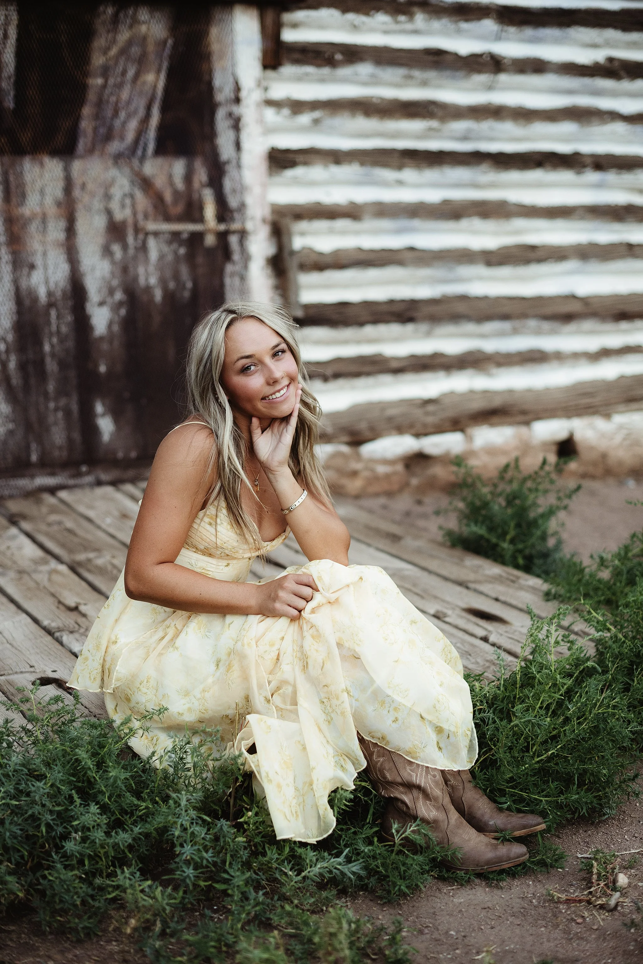 A young woman with long blonde hair, wearing a yellow dress and cowboy boots, sitting on wooden porch steps in front of a rustic wooden barn with whitewashed logs and a rusty door, smiling at the camera.