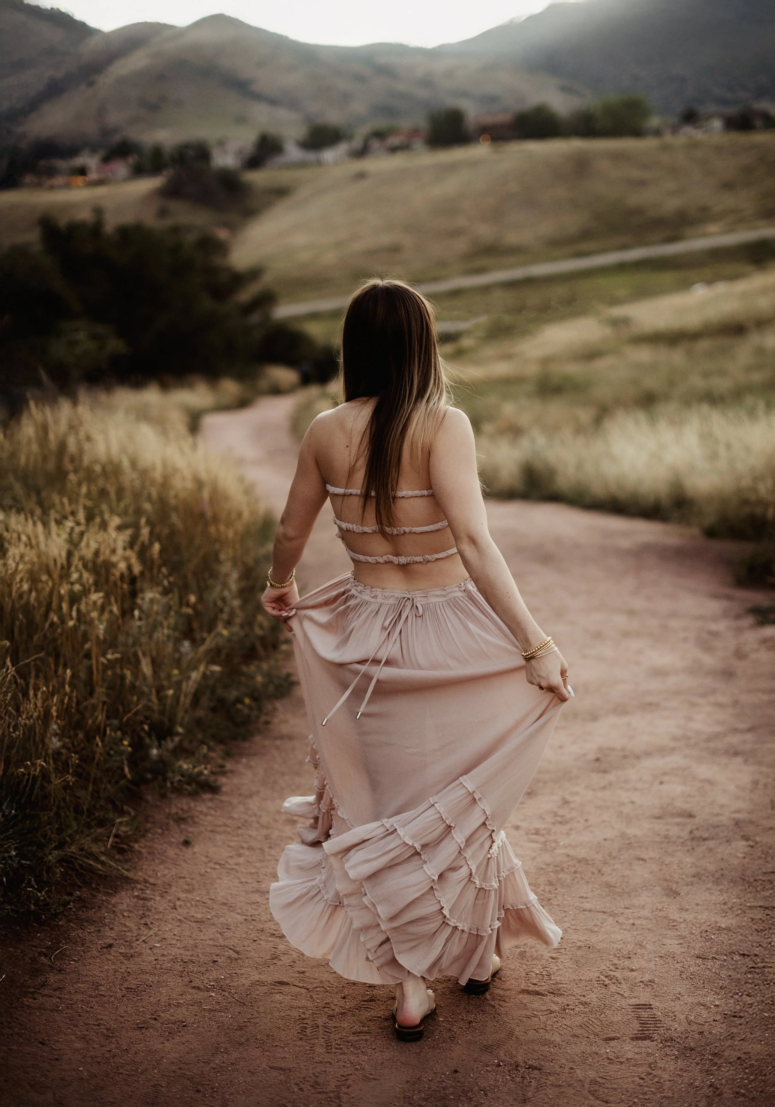 A woman in a flowing pink dress walking on a dirt path in a rural landscape with hills and small houses in the background.