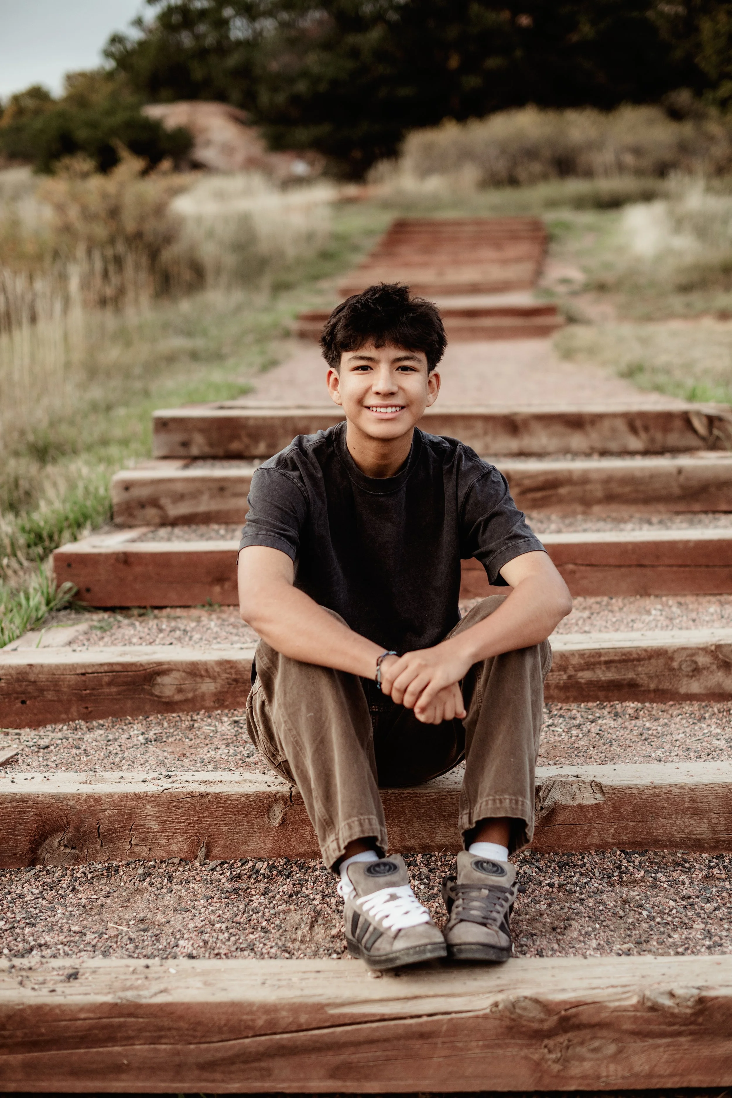 A young man with dark, curly hair, sitting on wooden outdoor steps, smiling, wearing a black t-shirt, brown cargo pants, and sneakers, with a natural landscape of trees, grass, and a wooden fence in the background.