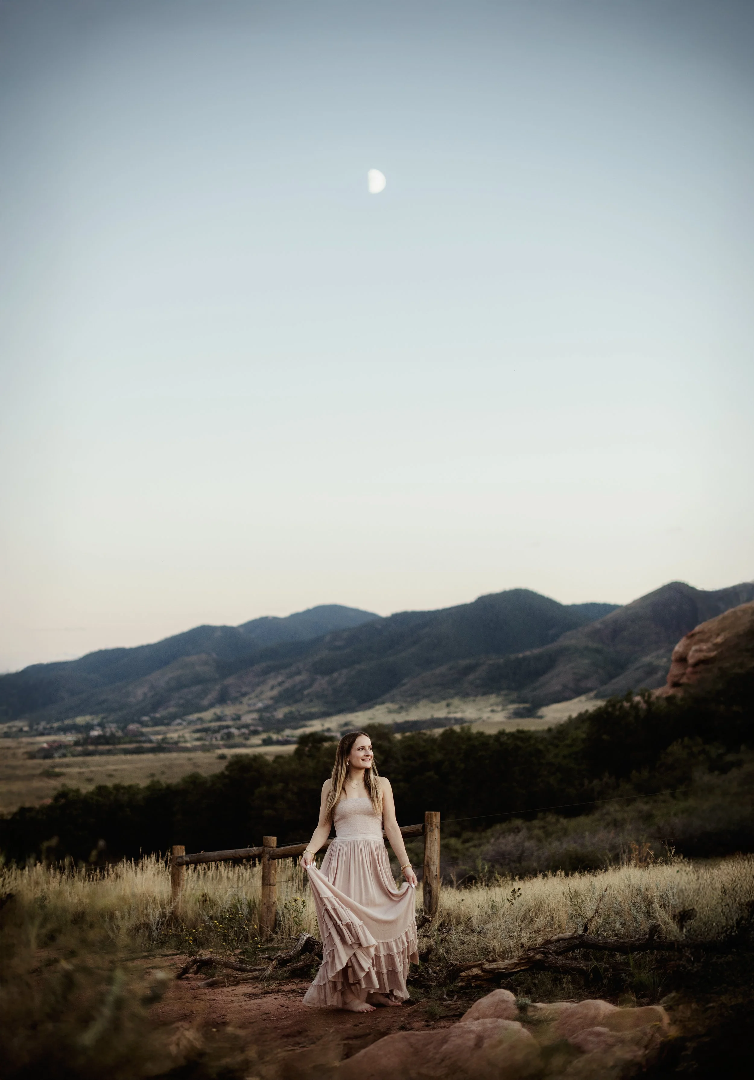 A woman in a long beige dress standing outdoors near a fence, with mountains and a moon in the sky during sunset.