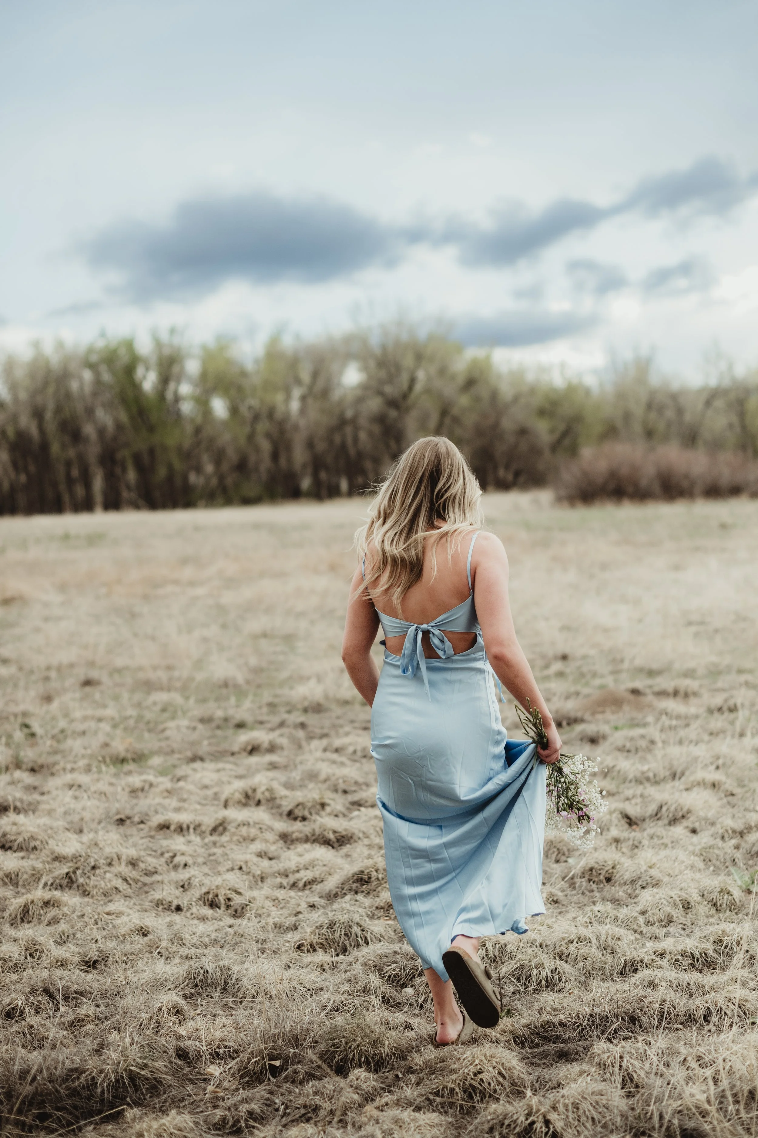 Woman in a light blue dress walking barefoot in a field, holding flowers, with trees and a cloudy sky in the background.