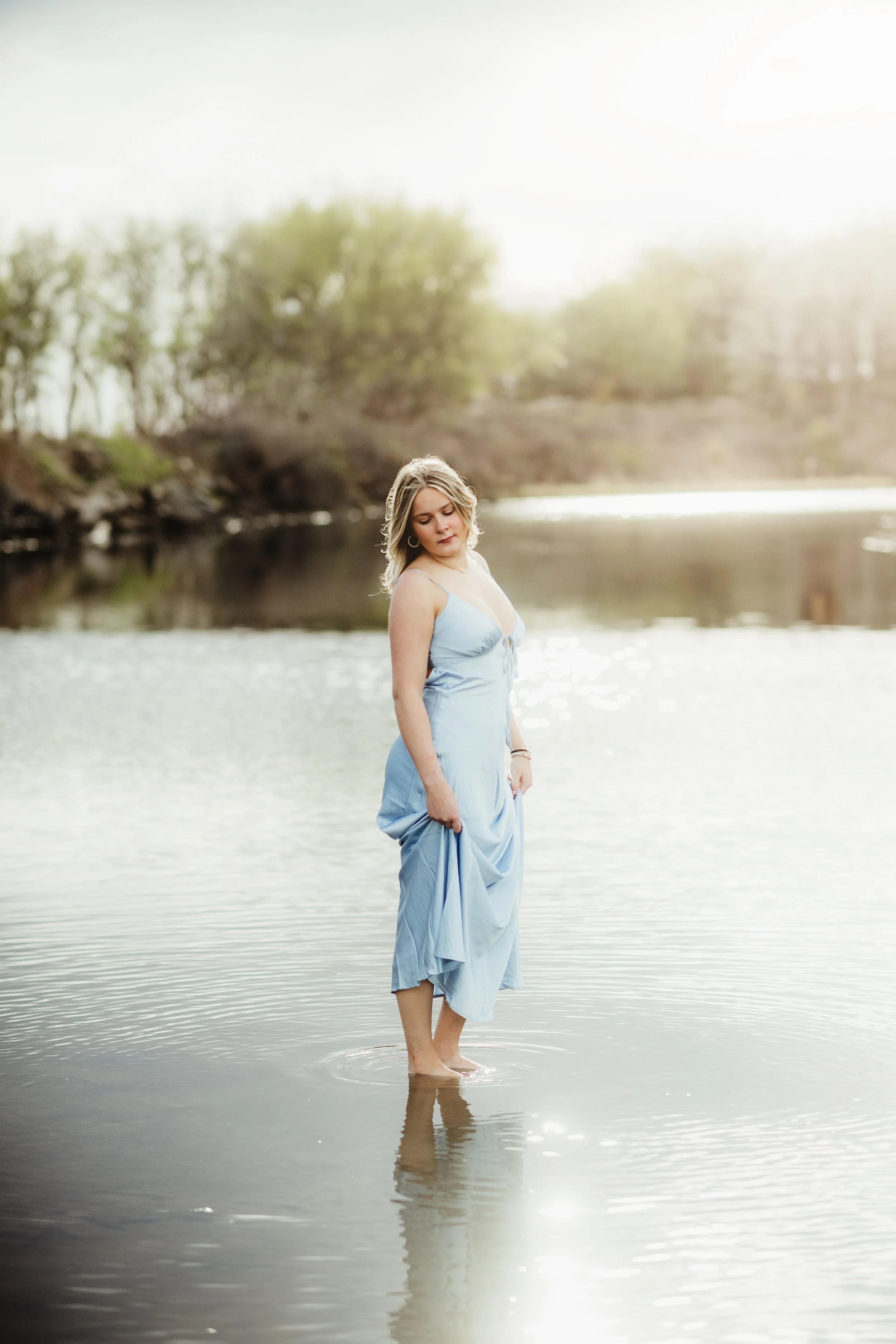 A young woman in a light blue dress is standing in shallow water by a lake or river, with her eyes closed and her head slightly tilted down. She is barefoot and holding the hem of her dress with one hand. The background features trees and a partly cl