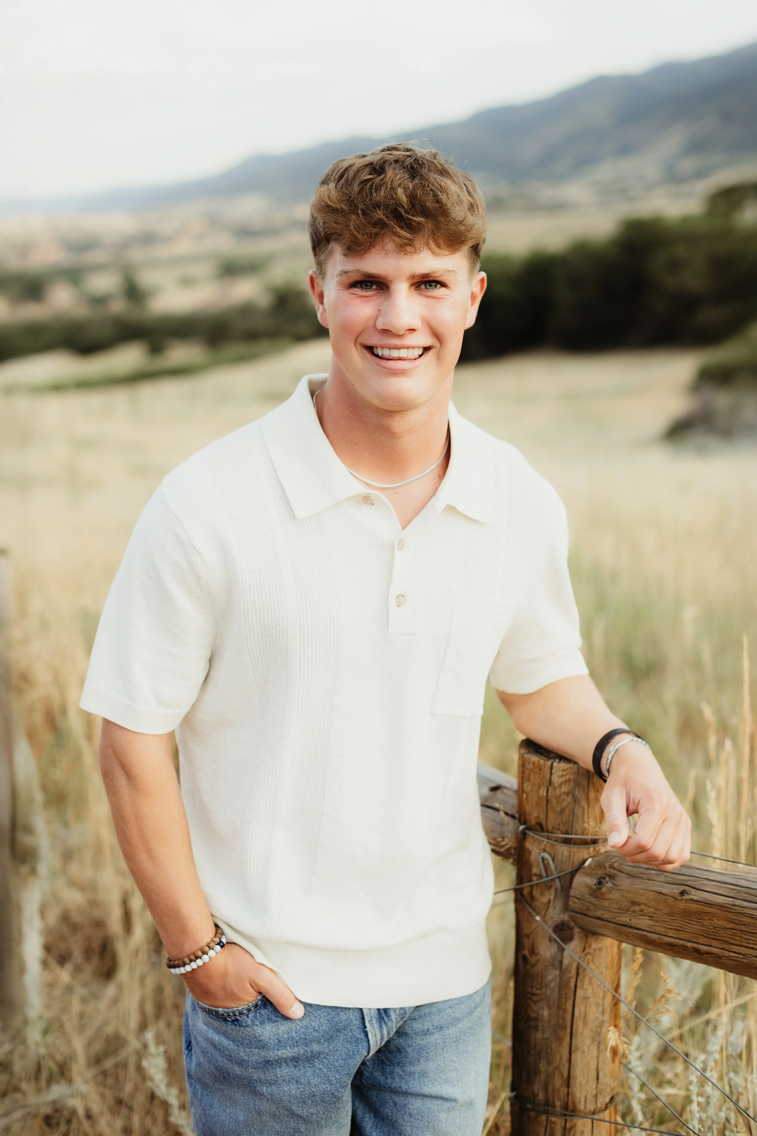 A young man with short, light brown hair and blue eyes smiling outdoors on a cloudy day, standing by a wooden fence in a rural area with fields and mountains in the background.
