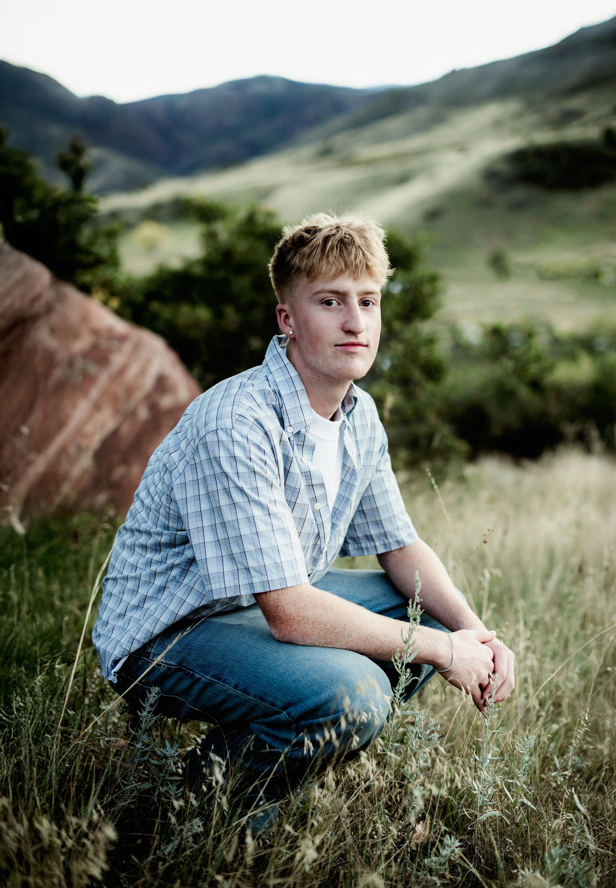 A young man with blond hair and freckles sitting in a grassy field with mountains in the background. He is wearing a blue checkered shirt and jeans, looking at the camera.