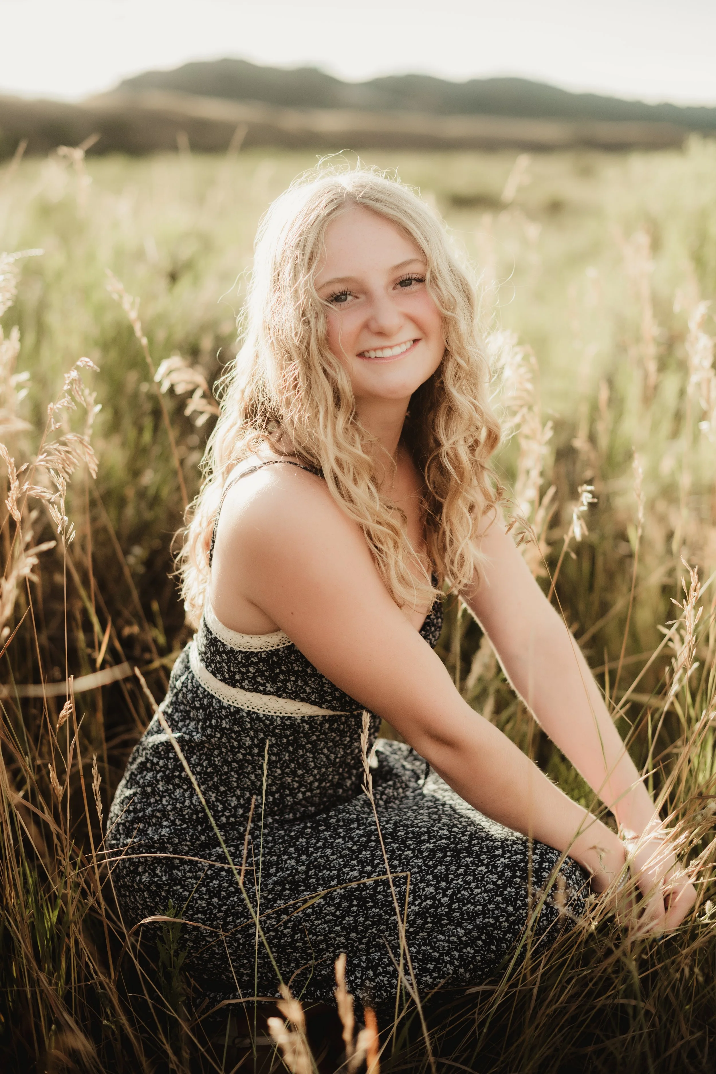 A young woman with curly blonde hair smiling while sitting in a field of tall grass with hills in the background, during golden hour.
