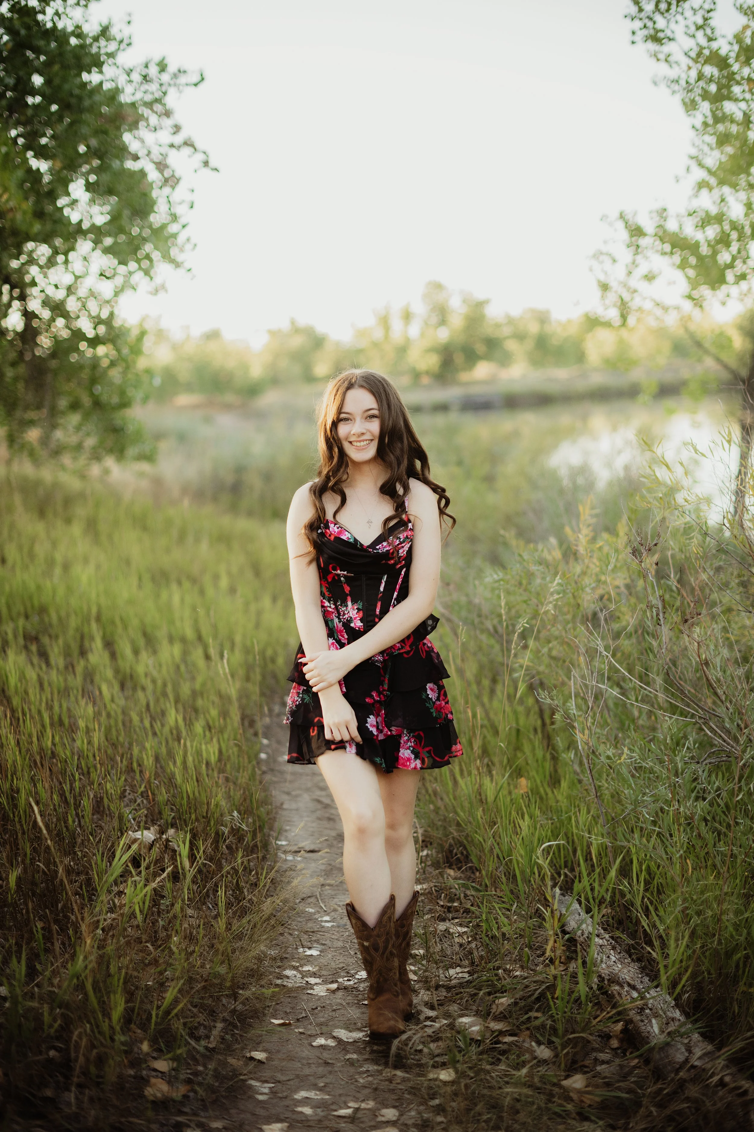 A young woman with long, wavy brown hair standing on a narrow dirt path in a grassy area with trees, smiling, wearing a black and pink floral dress, and cowboy boots in a natural outdoor setting during late afternoon or early evening.