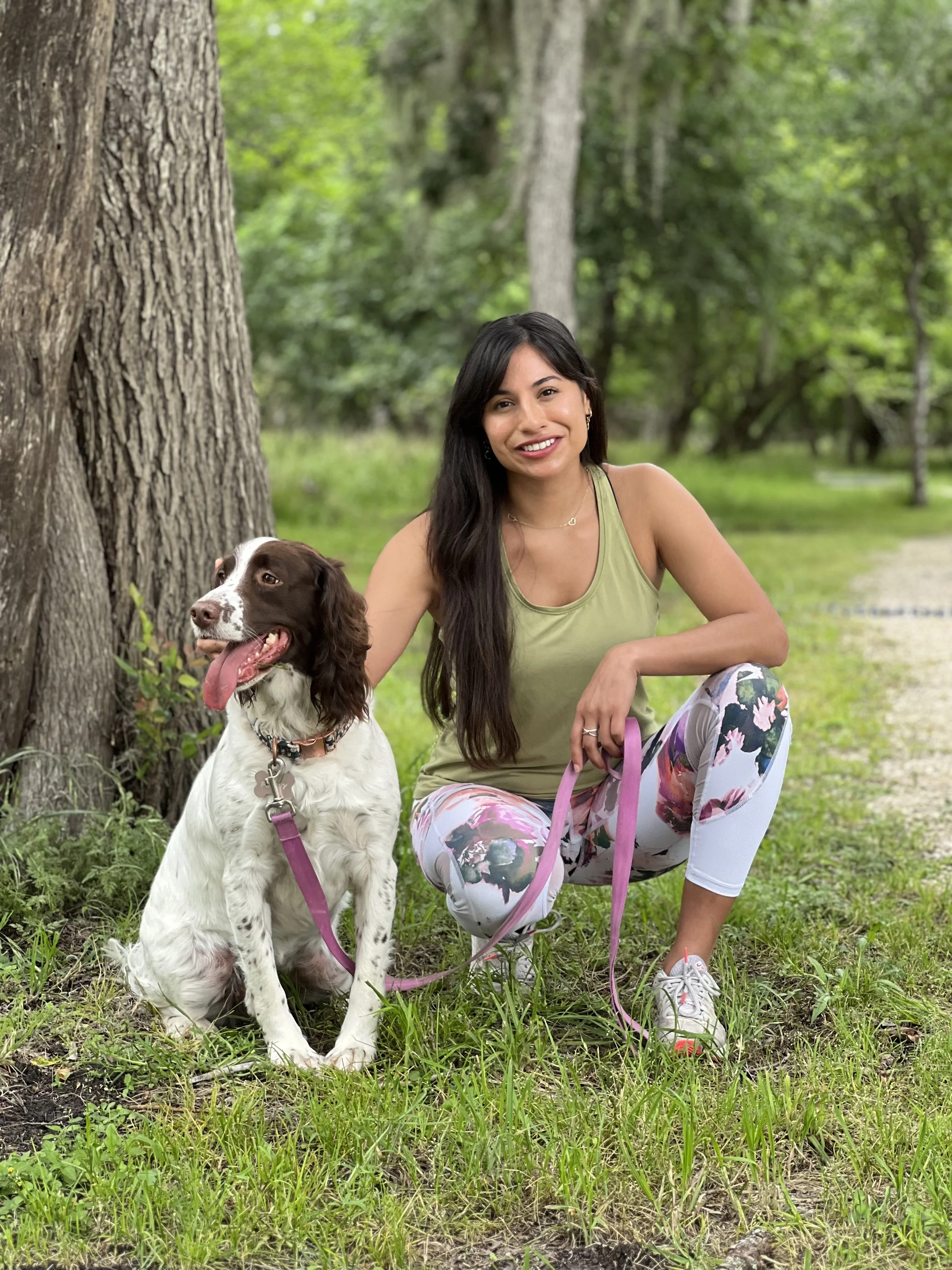 A woman kneeling on grass next to a brown and white dog with a pink leash, set against a background of trees.