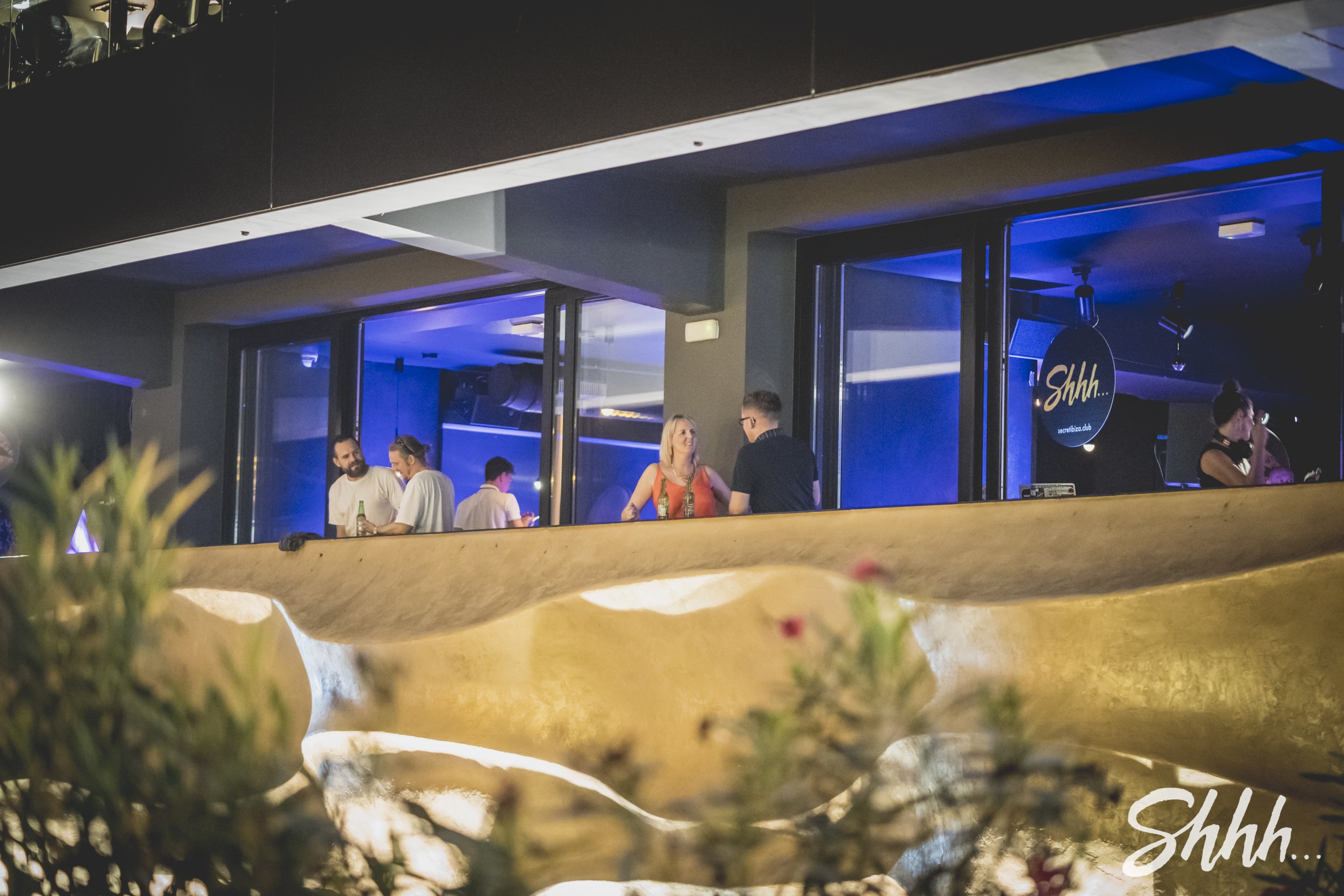 People socializing inside a dimly lit bar or club with blue lighting, visible through large windows, with some standing and talking, and one woman near the window drinking, background with a DJ booth or stage area, and a sign that says "Shhh...".