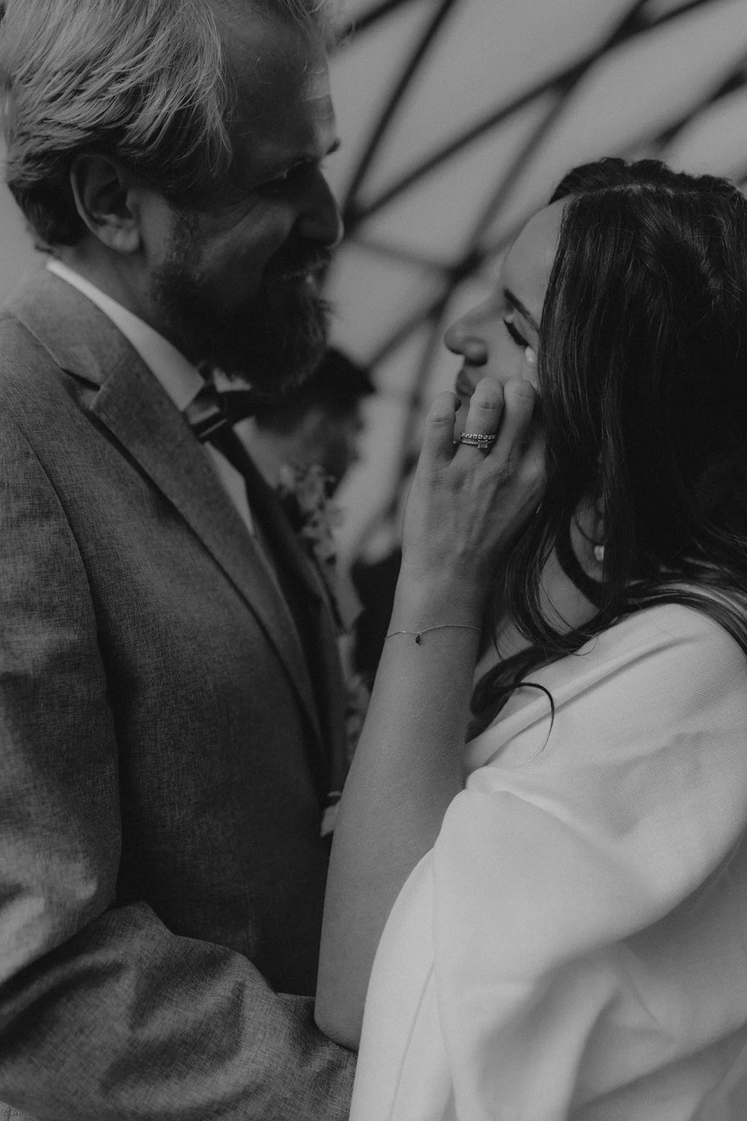 Black and white photo of a couple in formal attire embracing and smiling at each other, with grid-like ceiling structure in the background.