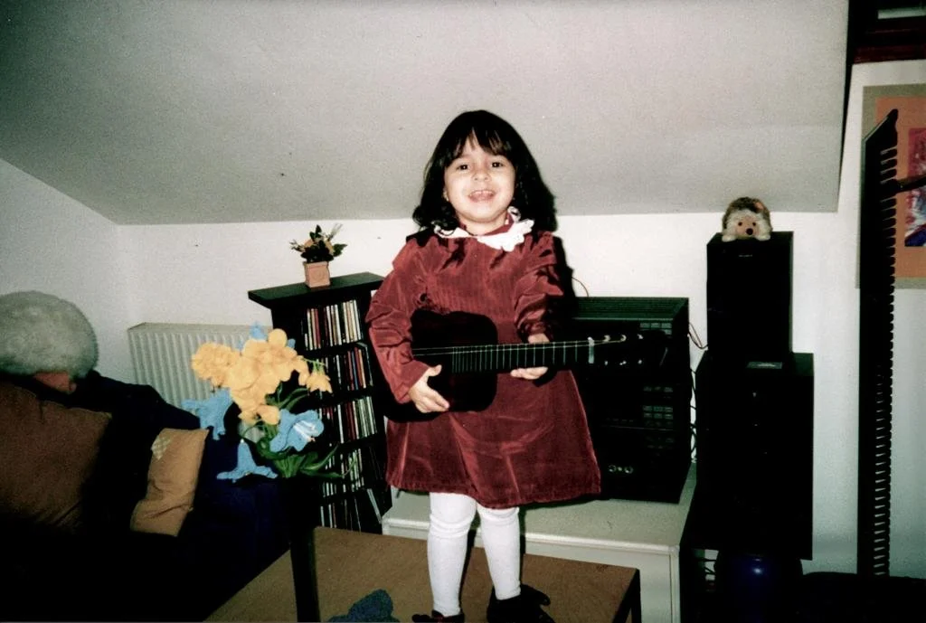 Ein kleines Mädchen in einem roten Kleid spielt Gitarre in einem Wohnzimmer. Im Hintergrund befindet sich ein Regal mit CDs und Lautsprechern. Eine Vase mit gelben Blumen steht auf einem Tisch.
