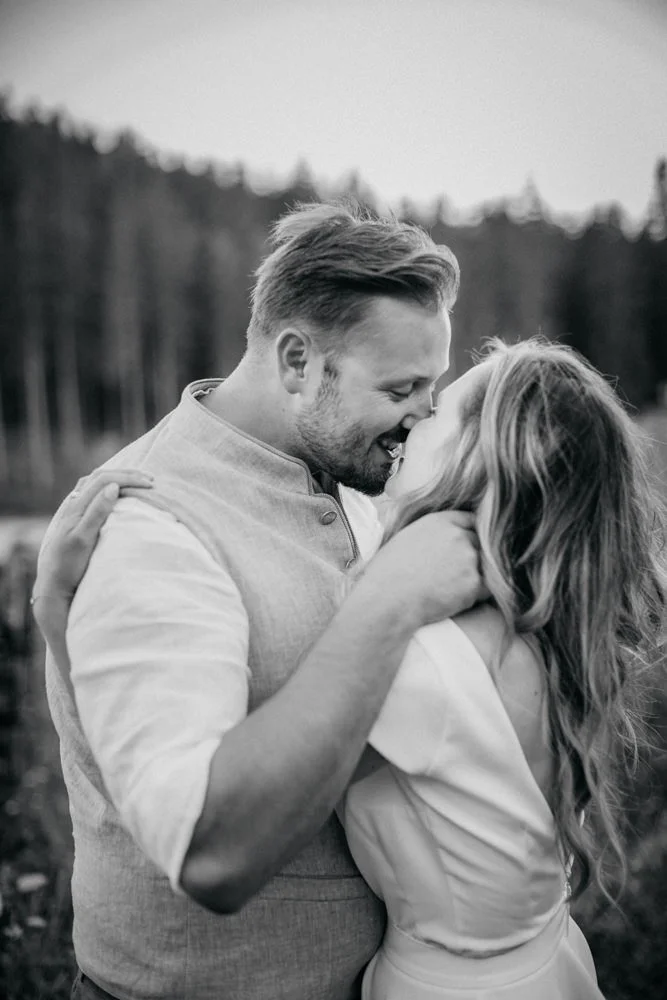 Ein Schwarz-Weiß-Foto eines Paares, das sich im Freien umarmt, die Gesichter dicht beieinander und lächelnd, im Hintergrund von Bäumen umgeben.