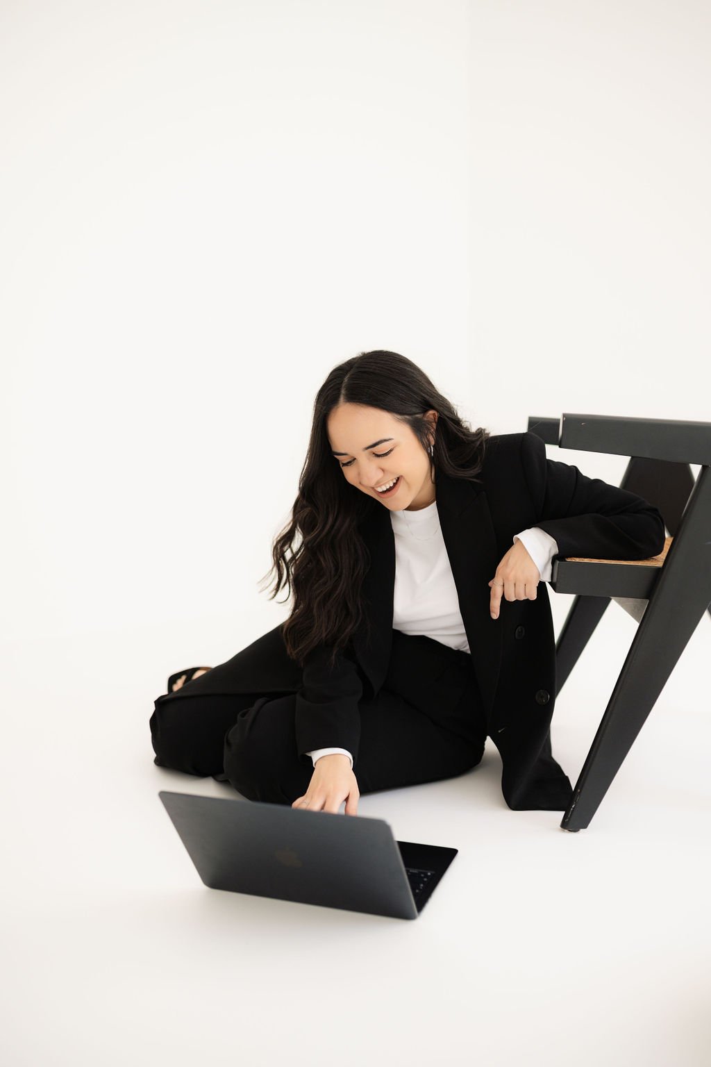 A woman in formal black suit and white shirt smiling and leaning on her knee, with a laptop on the floor in front of her and a chair turned upside down beside her, against a plain white background.