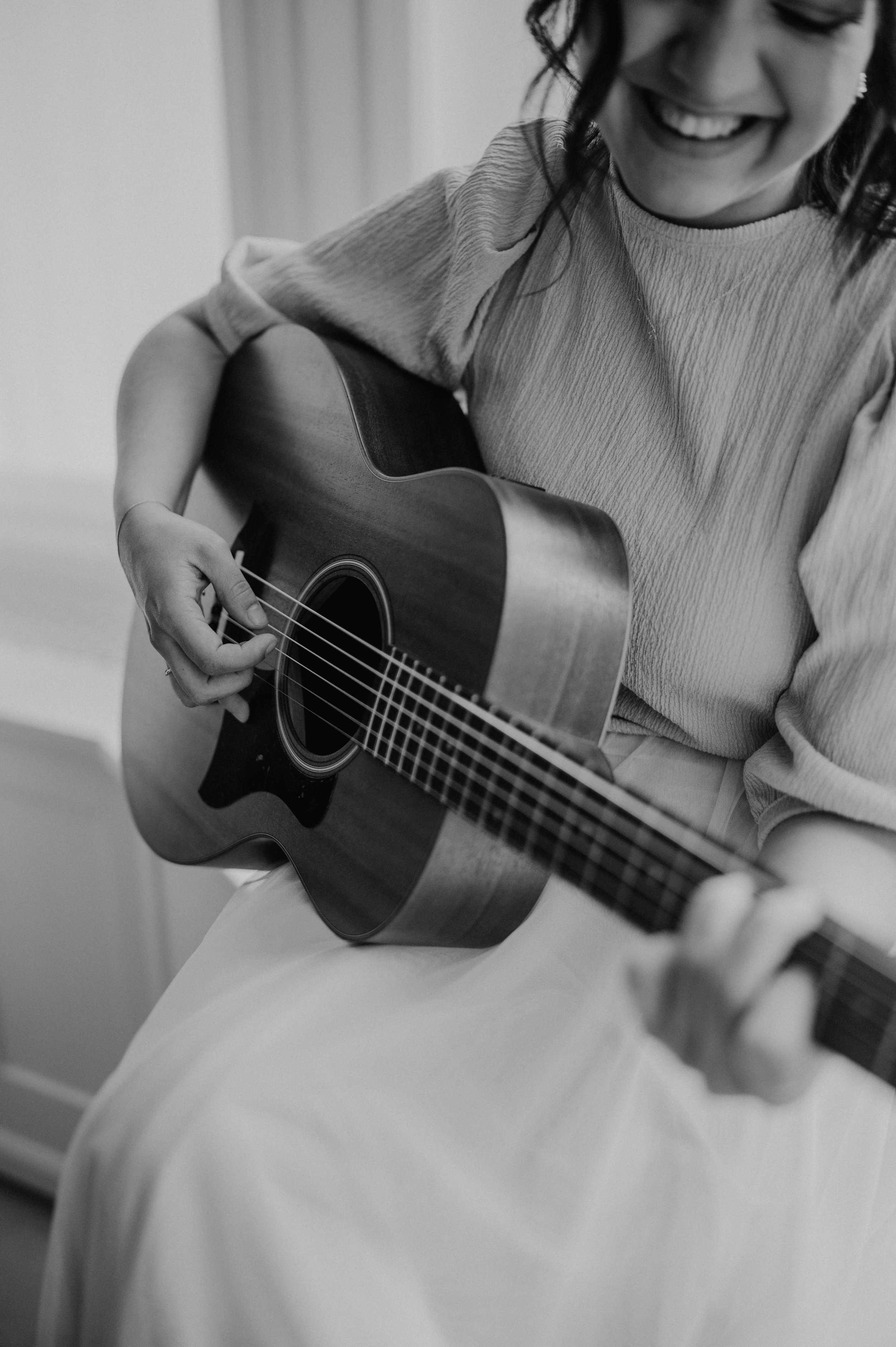 A woman smiling and playing an acoustic guitar in a black-and-white photograph.
