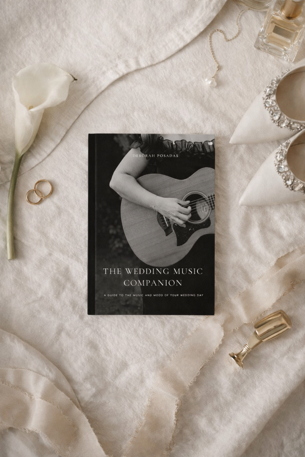 Ein Buch mit dem Titel „The Wedding Music Companion“ von Deborah Posadas, das auf einer weißen Stoffunterlage liegt und von Hochzeitsaccessoires umgeben ist, darunter goldene Ringe, eine weiße Calla-Lilie, eine Perlenkette, verzierte Schuhe mit Strasssteinen, ein kleines Parfümfläschchen und ein goldenes Schnapsglas.