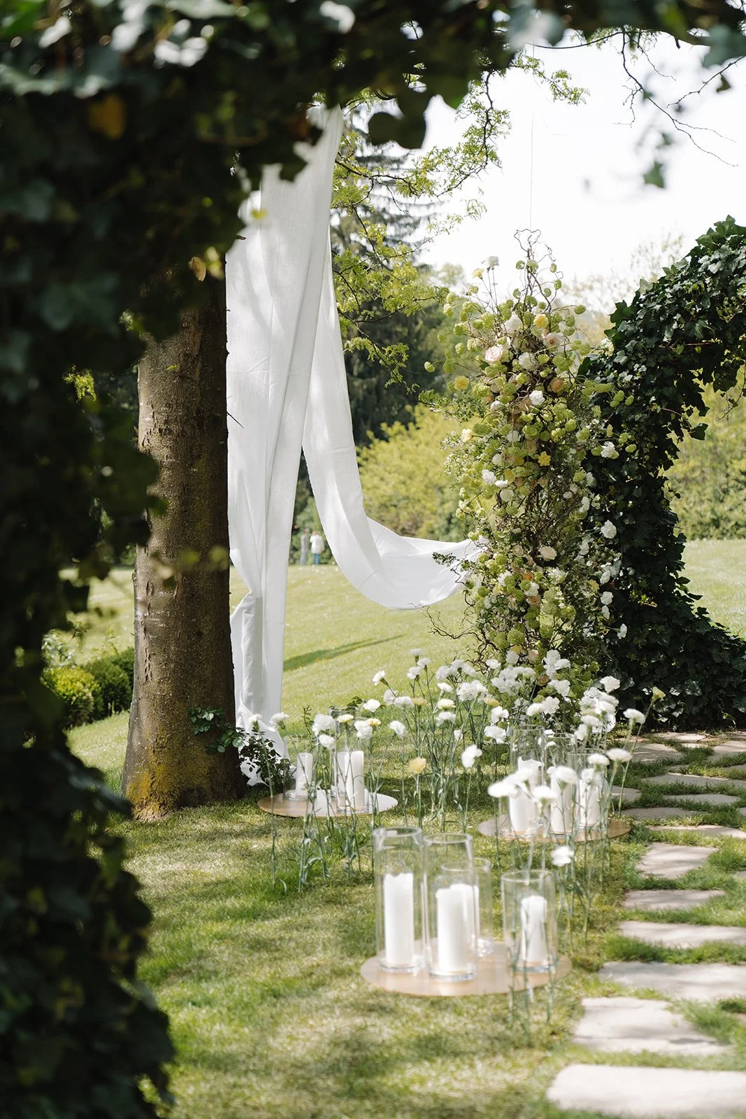 Outdoor wedding setup with a white drape hanging between trees, surrounded by green foliage and flowers, with candles and glass jars with white flowers on the ground.
