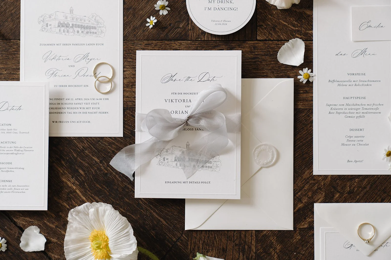Wedding invitation suite on a wooden table, includes white cards with elegant black script, two gold wedding rings, white flowers, and petal decorations.