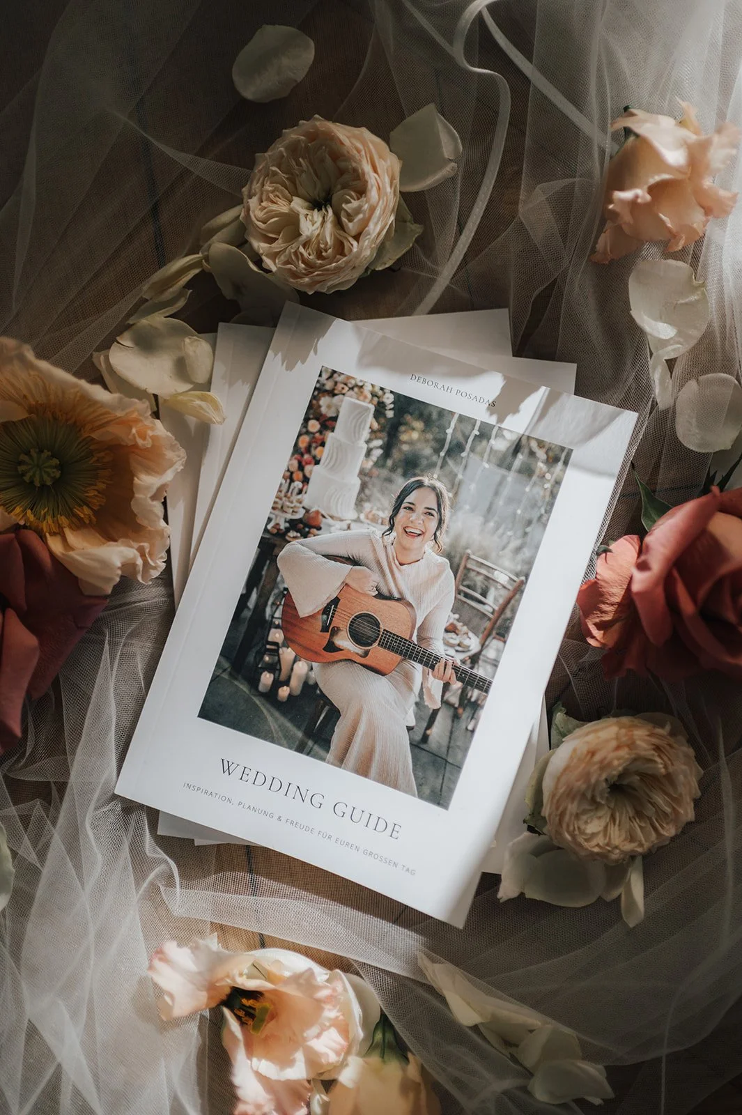 Ein Hochzeitsratgeber mit einer lächelnden, Gitarre spielenden Frau, umgeben von verstreuten Blumen und durchsichtigem Stoff.