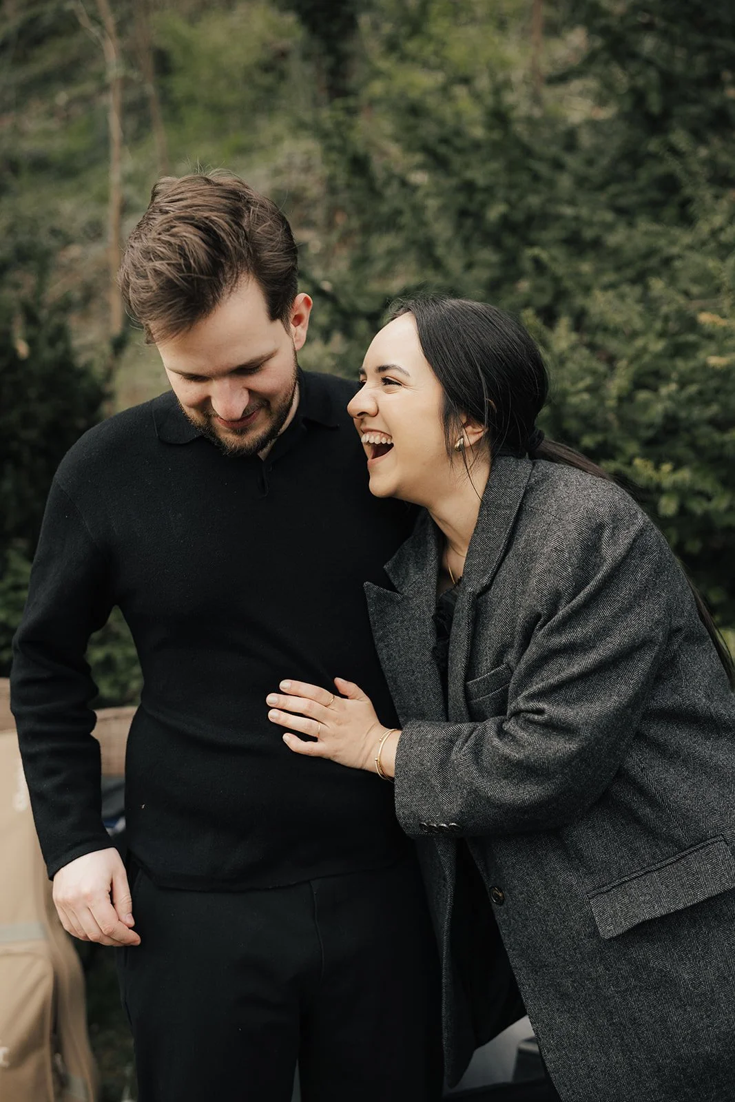 A smiling woman in a gray coat playfully interacts with a man in a black sweater in a park setting.