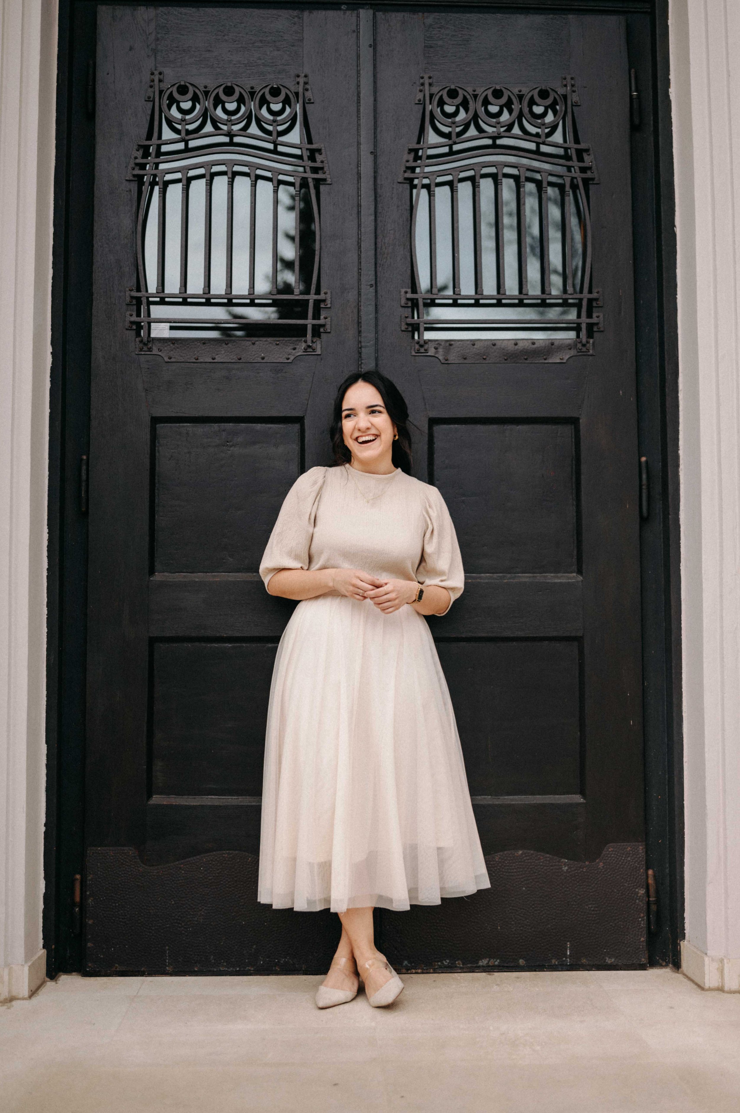 A woman in a cream-colored dress with puffed sleeves, standing in front of a large black wooden door with decorative metal window grilles, smiling.
