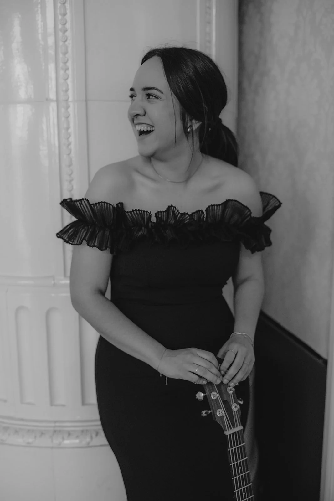 Eine lächelnde Frau mit nach hinten gebundenen dunklen Haaren, die ein schulterfreies Kleid mit Rüschen trägt, eine Gitarre in der Hand hält und in einem Raum mit gemusterten Wänden steht.